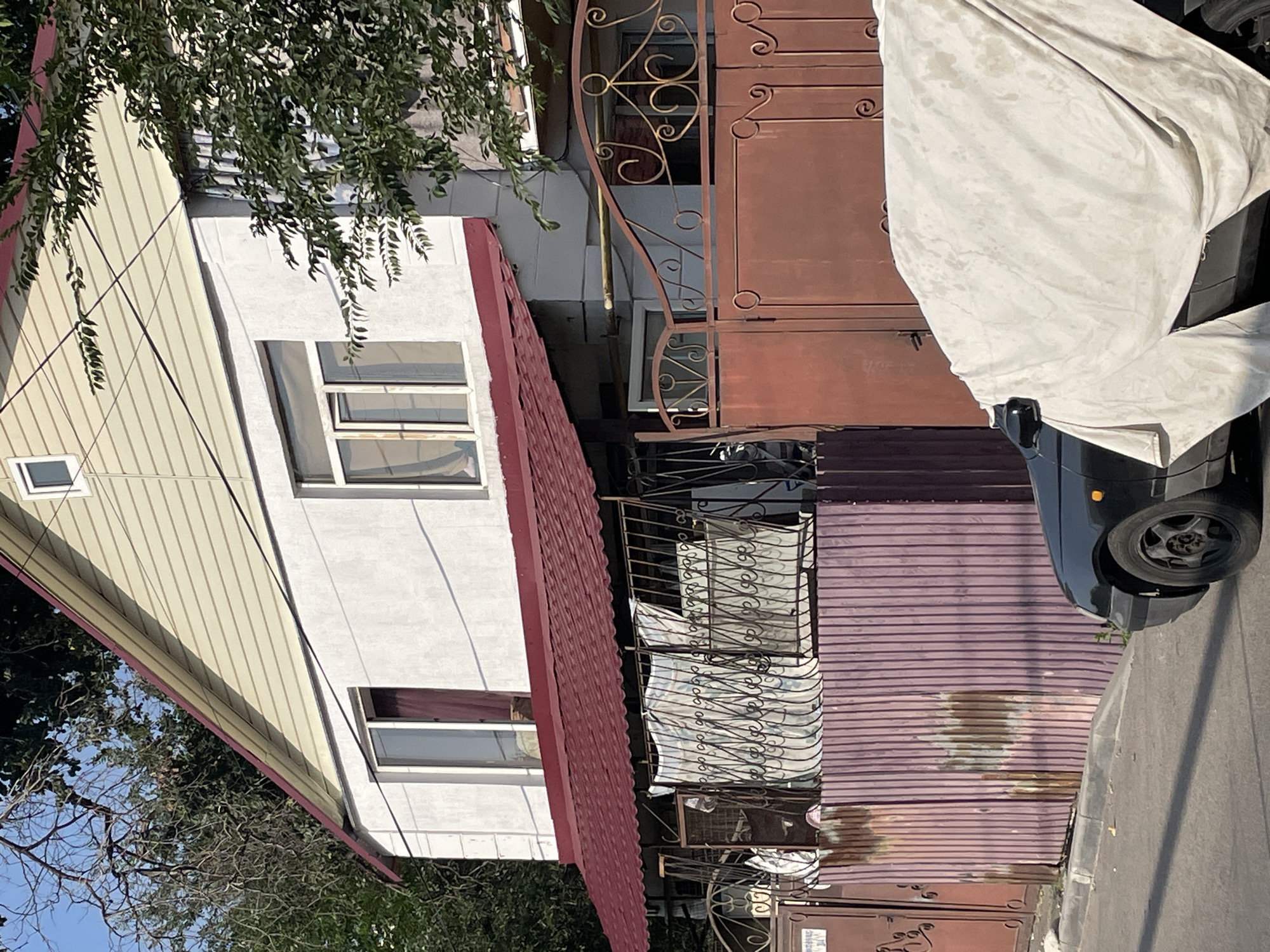 A two-story house with beige siding on the upper level and white stucco on the lower level is shown. The house has a red metal roof that extends over a covered porch area with decorative black metal fencing. Behind the fencing, there are some draped fabrics. A purple corrugated metal fence runs along the front of the property, with a section of red metal gate visible on the right. A black car, partially covered with a light-colored tarp, is parked in front of the fence. Trees and foliage are visible around the house and on the right side of the image. The scene appears to be a residential street in Almaty, Kazakhstan.