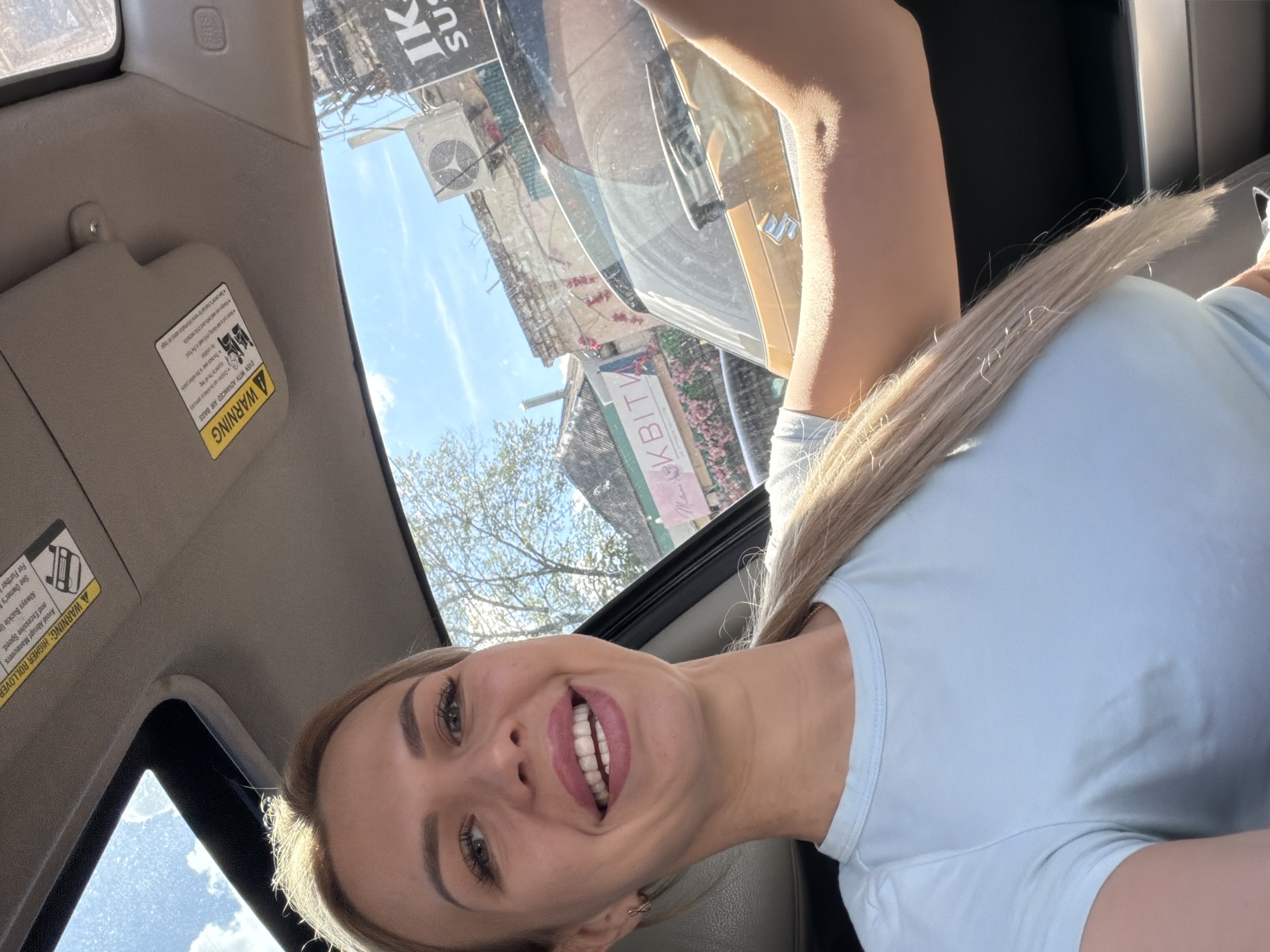 A woman with blonde hair and makeup is taking a selfie in a car. She is smiling and wearing a light blue t-shirt. The car has a sunroof open, and the sky is blue with some clouds. Outside the car, buildings and trees are visible. One building has signs with Cyrillic text, including "IK" and "SUSHI". An air conditioning unit is mounted on the side of a building. The overall scene suggests a sunny day in a city.