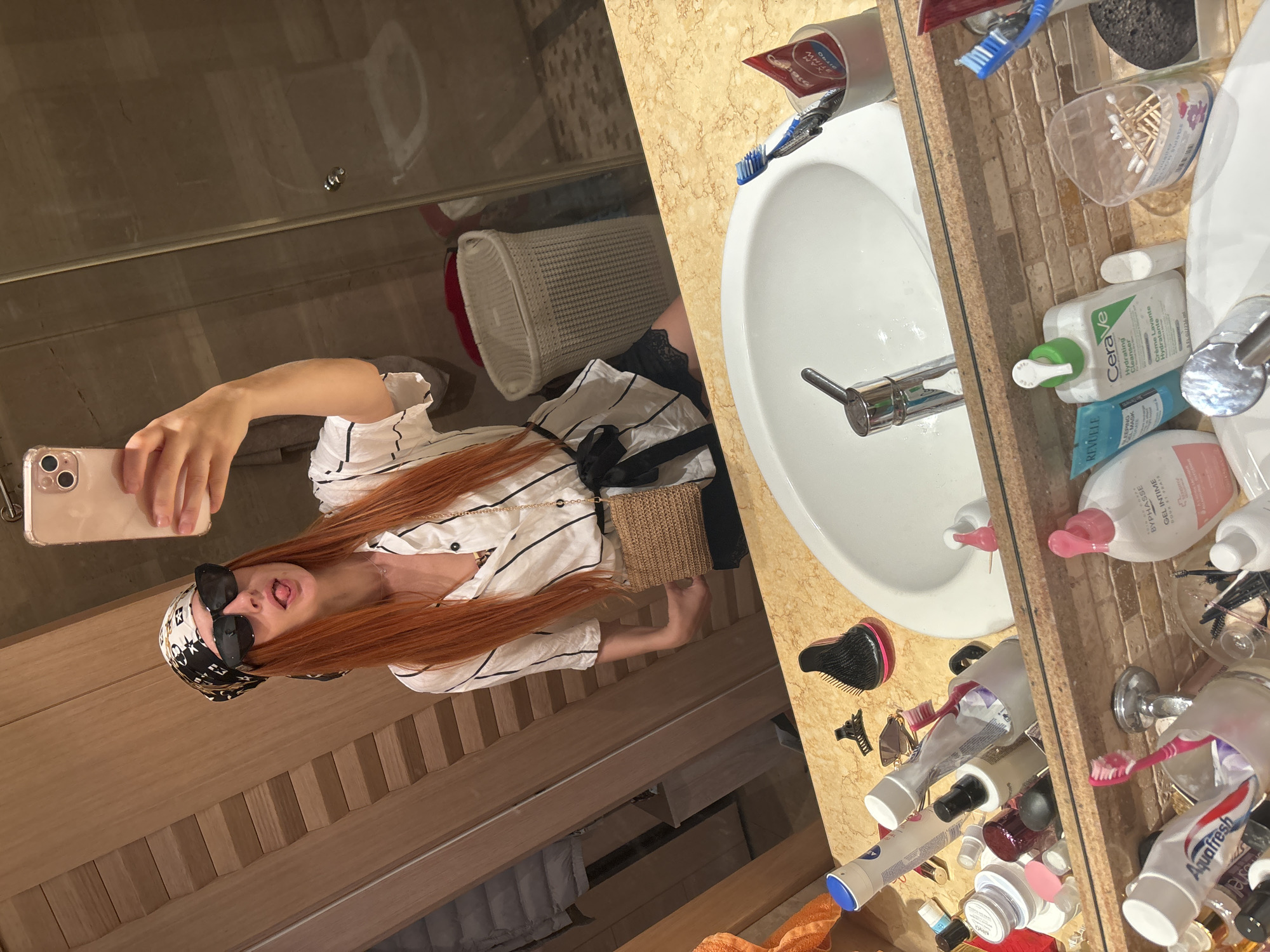 In a bathroom with a marble countertop and wooden cabinetry, a person with long, red hair and sunglasses is taking a mirror selfie. They are wearing a white and black striped shirt, a black and white bandana, and a small woven bag. Their tongue is sticking out, and they are holding a pink smartphone. The bathroom counter is cluttered with various toiletries, including toothbrushes, toothpaste tubes (Aquafresh, Colgate), cotton swabs, cleansers (Cerave Hydrating Cleanser, Revuele), and other bottles and containers. A white woven basket is also visible in the reflection. The scene appears to be indoors, possibly in the morning or evening, given the lighting. The overall mood is playful and casual.