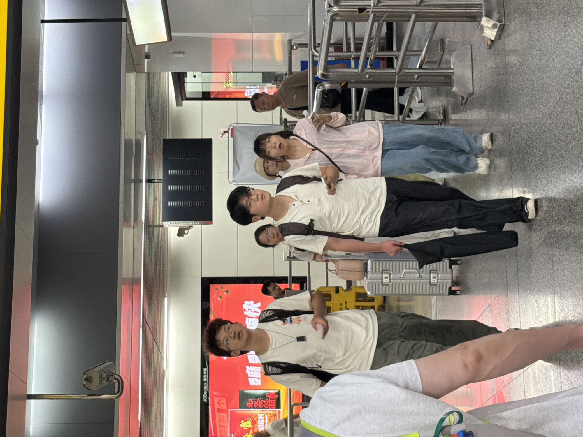 The image shows a group of people walking through an indoor public space, possibly a subway station or transit hub, in Jiulongpo District, China. A young man with curly hair and glasses, wearing a white t-shirt and a backpack, is in the foreground. To his right, another young man in a white t-shirt and dark pants, also with glasses and a backpack, is walking. Behind them, a woman in a pink jacket and blue jeans looks surprised, and a man in a brown shirt walks past. Several pieces of luggage, including a silver suitcase, are visible. The environment features a tiled floor, metal railings, and illuminated digital screens in the background. A security camera is mounted on the ceiling.
