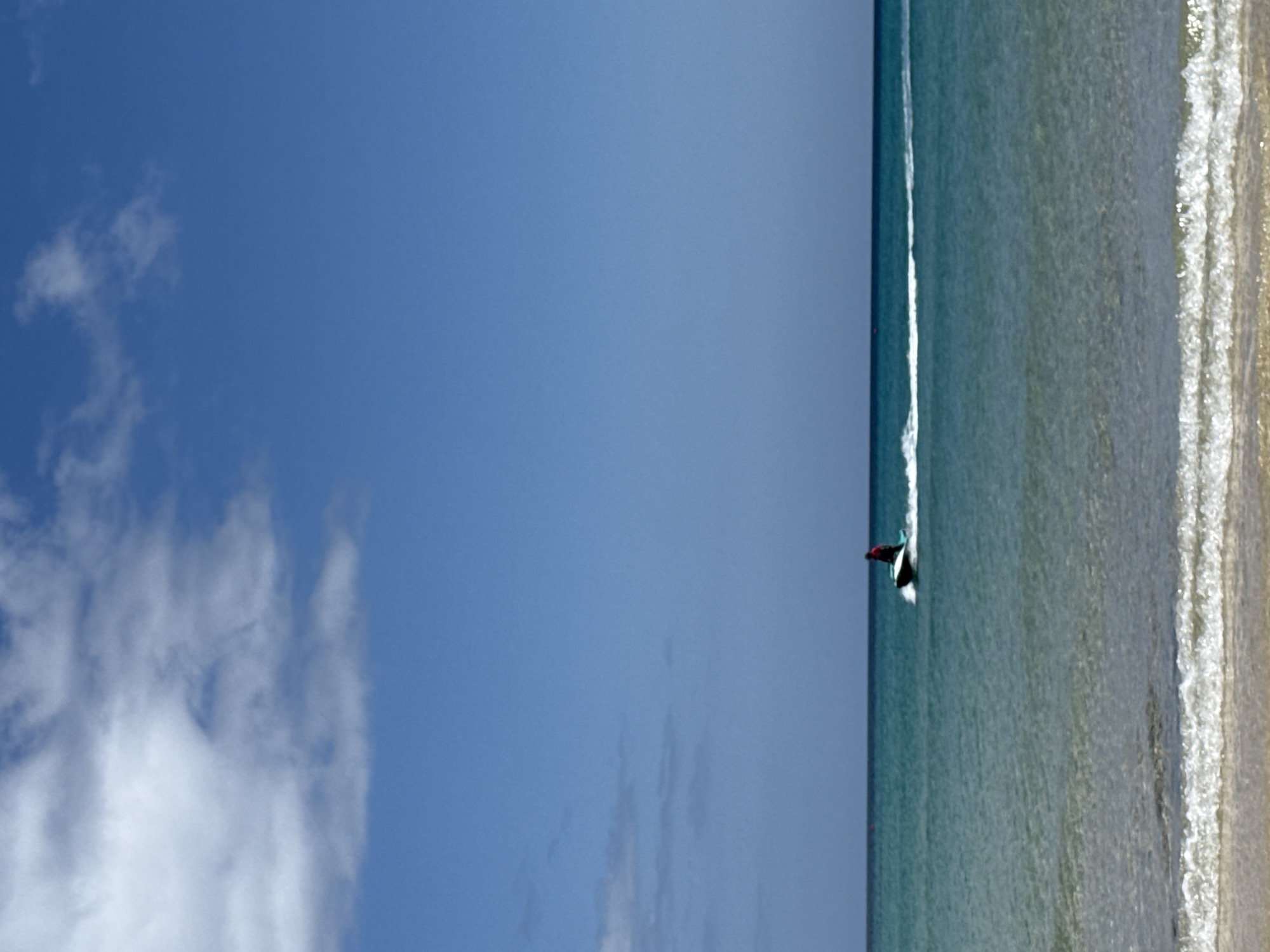 A bright, daytime image captures a serene coastal scene under a clear blue sky. The top two-thirds of the frame are dominated by the expanse of the sky, featuring scattered, wispy white clouds that are more concentrated towards the upper left. The bottom third of the image depicts calm ocean waters meeting a sandy shore.
In the ocean, a person is visible on a dark surfboard, appearing to be surfing or paddleboarding near the shoreline. The individual is partially obscured, wearing what appears to be a dark or red top. A thin line of white foamy water trails behind the surfboard, indicating movement. The water is a clear, light blue-green hue, with small, gentle waves breaking into white foam as they approach the sandy beach on the right side of the frame. The beach itself is a light, sandy strip.
The scene suggests leisure and outdoor activity on a sunny day. The weather is clear and bright, consistent with a pleasant beach environment. There is no visible text in the image.