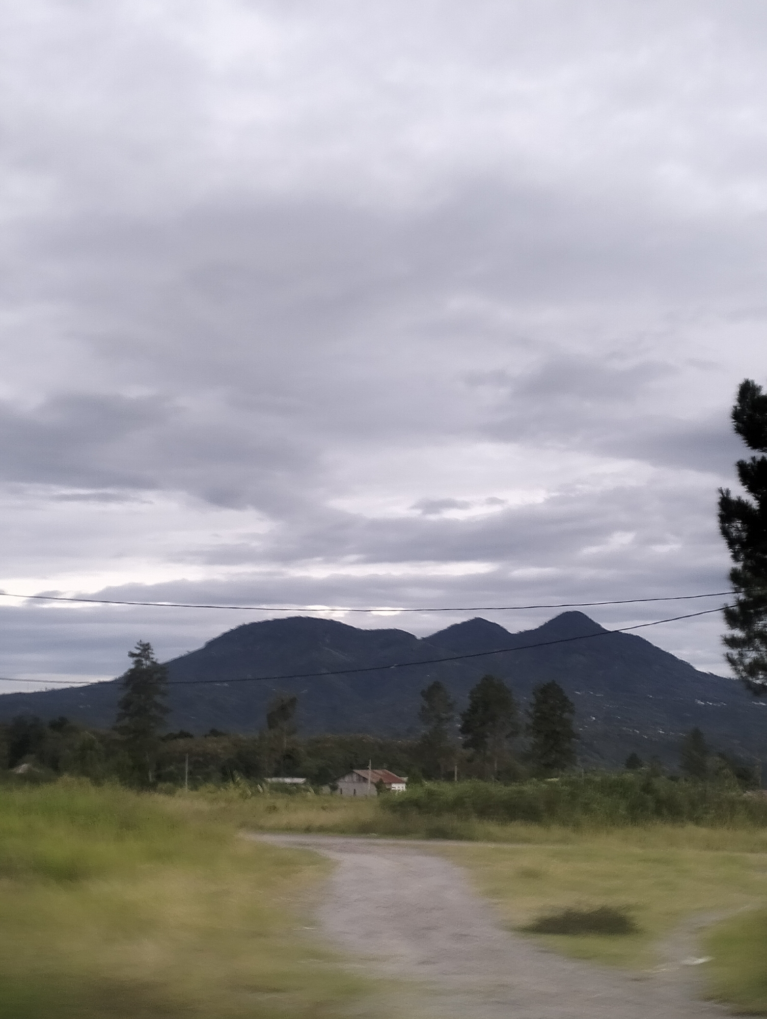 The image depicts a landscape scene with rolling hills or mountains in the background under a cloudy sky. In the foreground, a dirt road or path curves through a grassy area. There are scattered trees and some vegetation. A small building, possibly a house, is visible amongst the trees in the middle ground. Power lines are present, stretching across the scene horizontally in the mid-background. The overall lighting suggests it is daytime, likely overcast or early morning/late evening. The location is identified as Karangrejo, Indonesia.