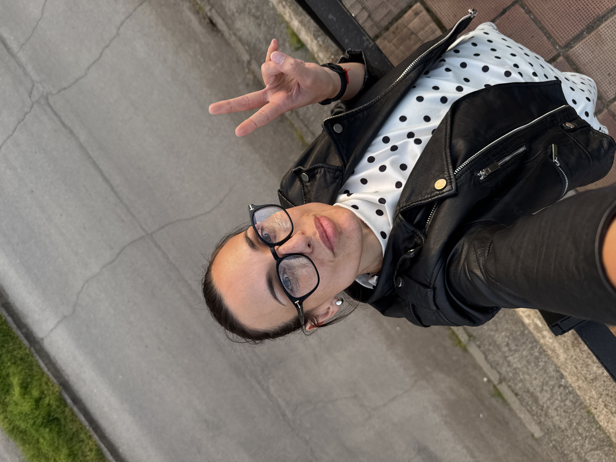 An overhead, slightly tilted selfie captures a woman outdoors in Pleven, Bulgaria. She looks directly at the camera with a neutral expression. Her dark hair is pulled back, and she wears black-rimmed glasses, which have a small "T" visible on the side of the left frame. She is dressed in a black leather jacket, featuring zippers and snap buttons, worn over a white blouse with black polka dots. On her right wrist, she wears a red string bracelet and a black watch. Her right hand is raised, making a two-finger gesture, similar to a peace or victory sign, with her index and middle fingers extended. A silver stud earring is visible in her left ear.
The background shows a paved surface, likely concrete or asphalt, with several cracks. To the left, a narrow strip of green grass borders the pavement. A dark railing or fence post is visible in the upper right, suggesting she might be standing next to a barrier or on an elevated path. The soft, even lighting indicates it is daytime, possibly late afternoon or early evening, with clear weather.