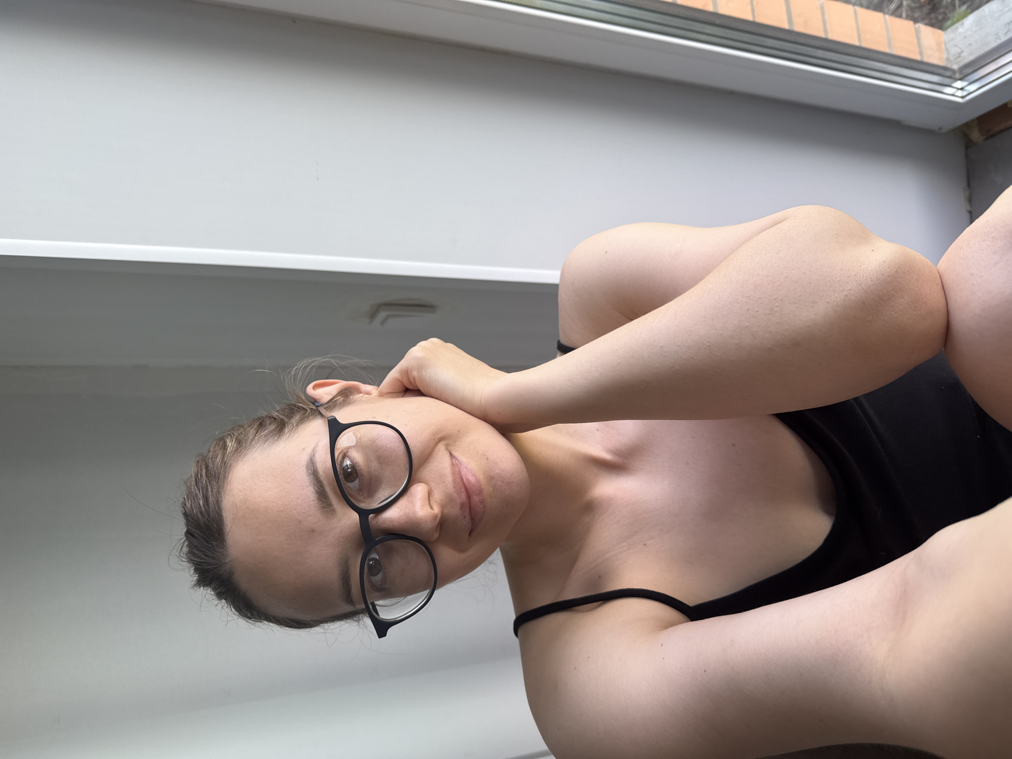 A woman with light skin, wearing black-rimmed glasses and a black tank top, poses indoors. Her hair is pulled back. She has her arm raised to her head. The location is unidentified; there is a window in the background and a light-colored wall. There is no evidence in the image to suggest this image was taken in Chaikovsky, Russia.