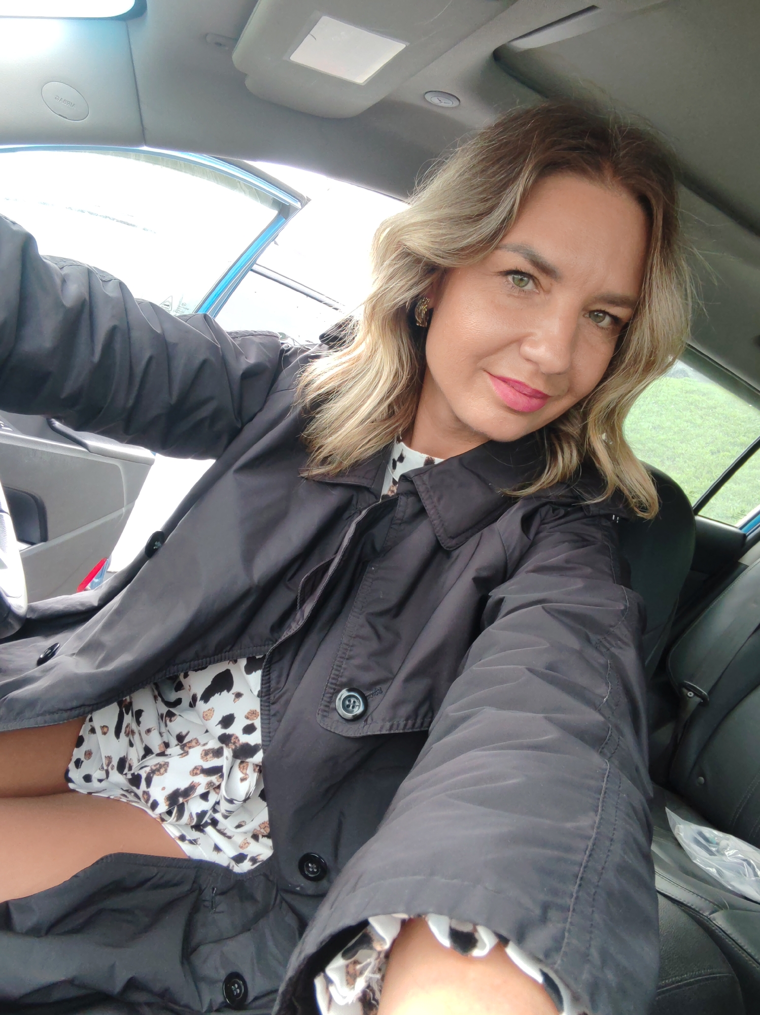 A woman is sitting in the driver's seat of a car, likely in a selfie pose. She is wearing a patterned dress and a dark-colored trench coat. The car's interior is visible, including the dashboard, steering wheel, and seats. The background seen through the car window appears to be a grassy area or field. There is no evidence in the image to support or contradict the location of Petropavlovsk-Kamchatsky, Russia.