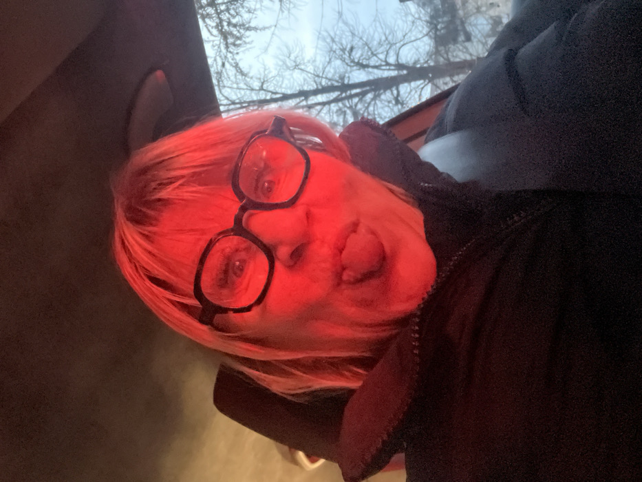 A person with short, blonde hair and large, black-rimmed glasses is illuminated by a red light. They are sticking out their tongue. The background includes blurred trees, possibly seen through a window. The person is wearing a dark-colored, possibly black, jacket. The image appears to be taken from a slightly upward angle.