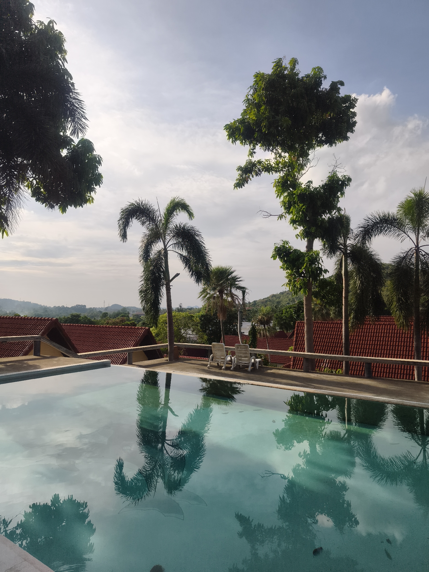 The image shows a swimming pool in บ้านบางรักษ์, Thailand. The pool is surrounded by trees and buildings with red tiled roofs. Reflections of the trees and sky are visible in the pool water. Two white lounge chairs are positioned near the pool's edge. In the distance, hills can be seen.