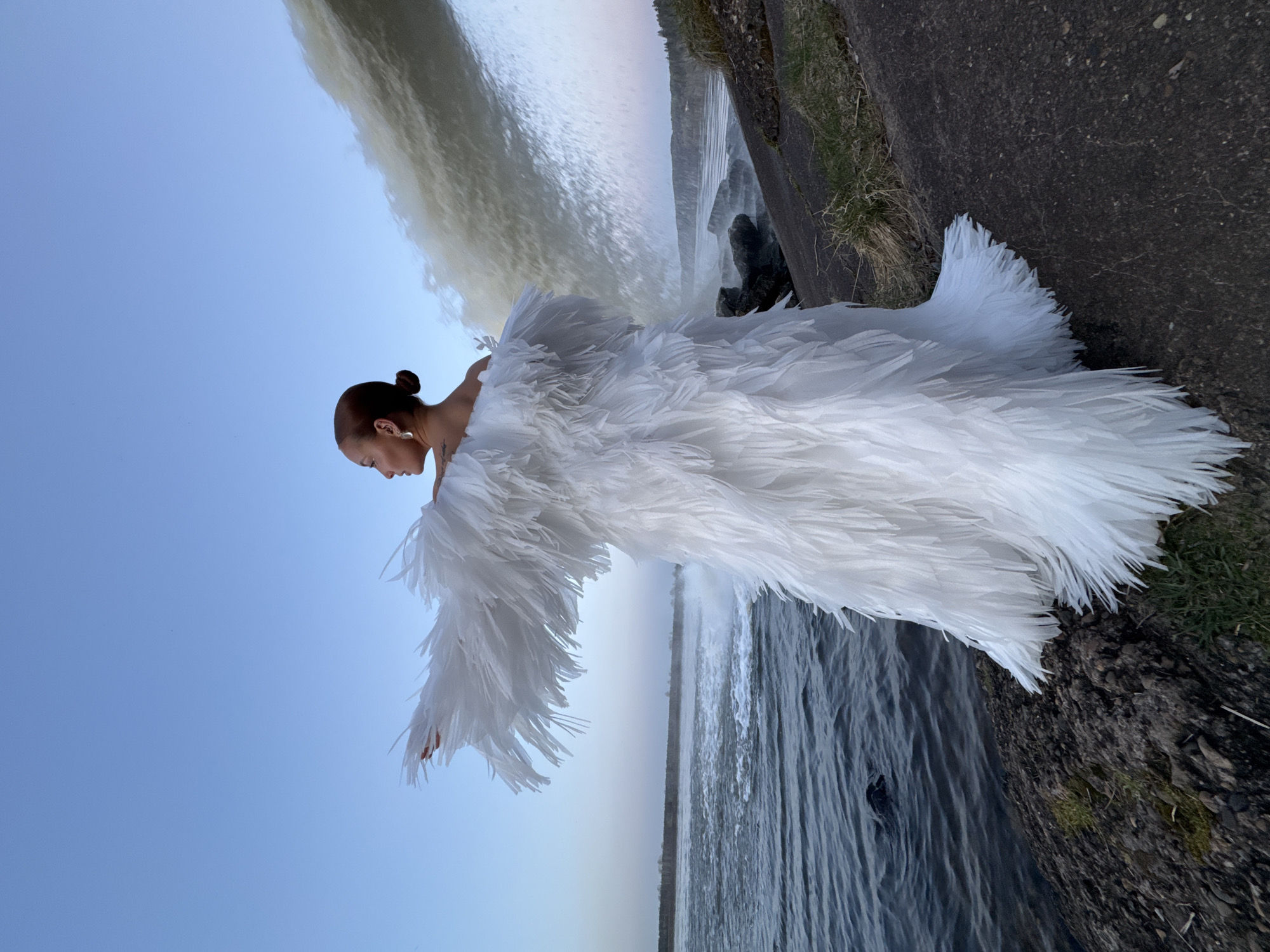 A rotated image shows a woman in a voluminous white feathered dress standing at the edge of a waterfall. The waterfall is to her right, cascading into a body of water below. The sky is pale blue in the upper left, gradually fading towards the horizon. To the woman's left, the waterfall has an earthen bank with grass and moss growth. The woman has her hair pulled back, and she appears to be looking down. The city of Noviy, Russia, cannot be confirmed as the location based solely on the visual information.
