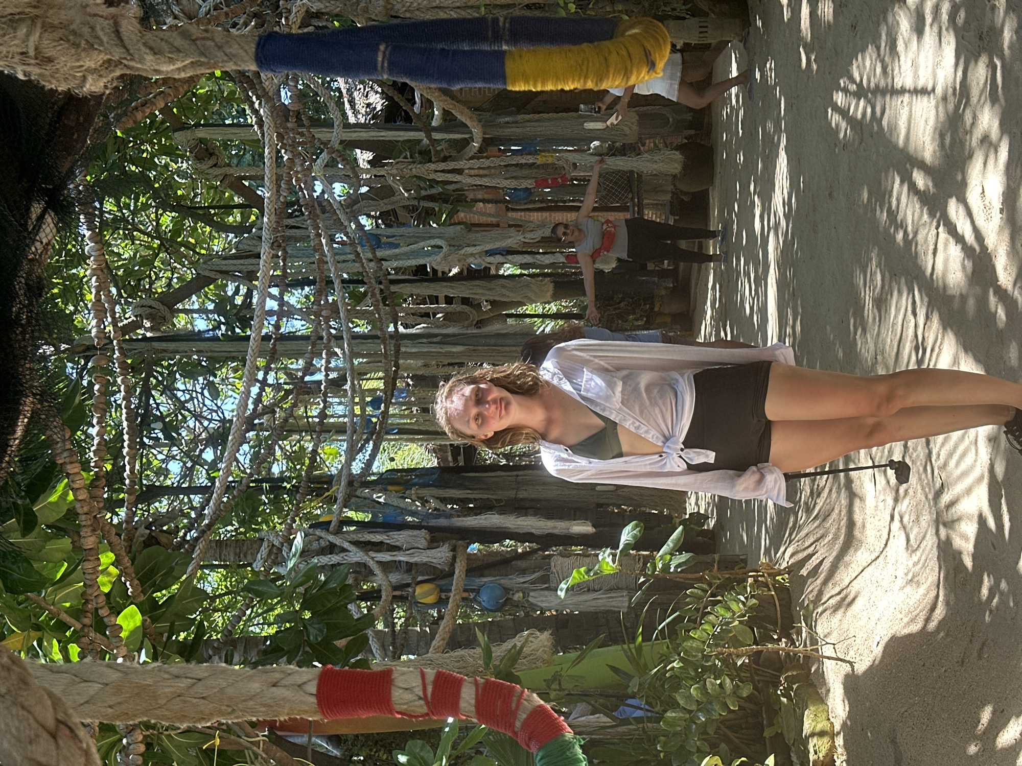 In Ko Pha-ngan District, Thailand, a young woman stands facing the camera on a sandy path lined with lush greenery. She wears a white unbuttoned shirt tied at the waist over a green bikini top and black shorts, with a golf club in her right hand. Behind her, a woman in a grey tank top and black pants has her arms outstretched, while another woman in white is taking a photo with her phone. The scene is outdoors, beneath a canopy of ropes, netting, and palm fronds, creating a dappled light effect on the sand. The overall impression is a tropical, possibly resort, environment.