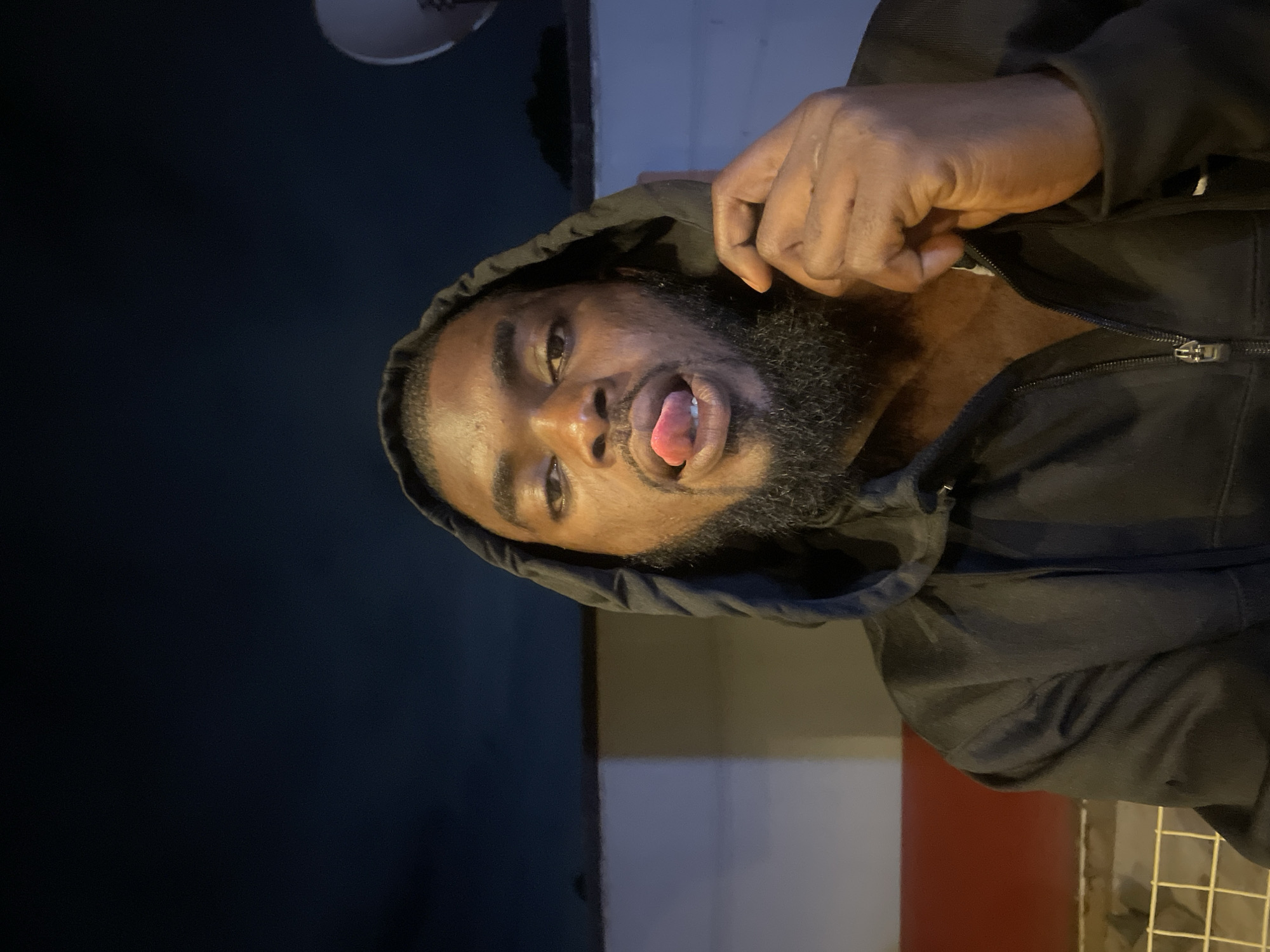 A man with dark skin, a beard, and a black hooded jacket is facing the camera with his tongue sticking out. The background shows a combination of dark and light elements, possibly indicating an outdoor setting at dusk or night. Based on general context, the location is likely within the city of Jos, Nigeria, although specific landmarks are not visible in the image.