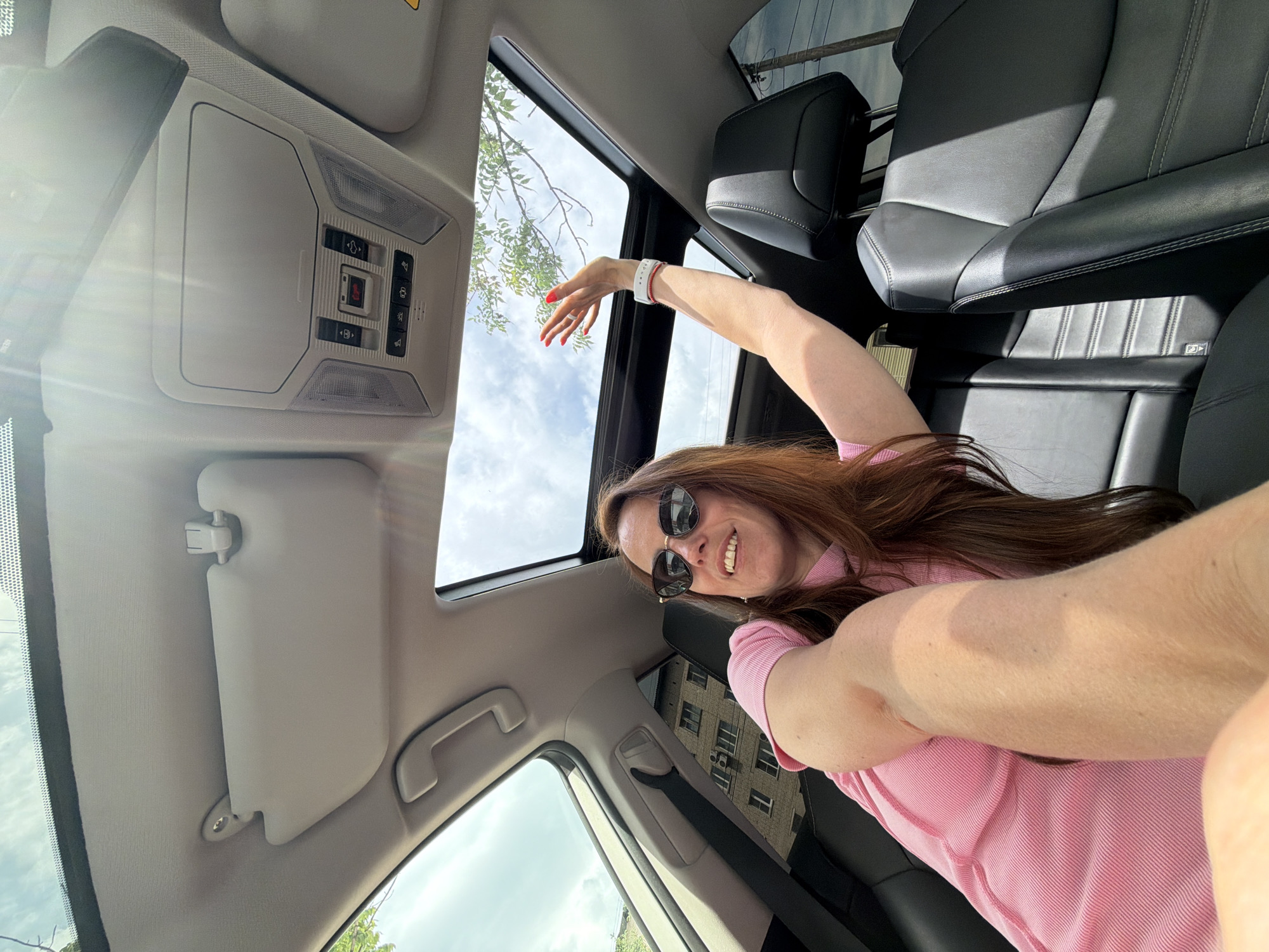 A woman with long auburn hair and sunglasses is sitting in the passenger seat of a car with the sunroof open. She is wearing a pink shirt and has her left arm extended upwards, reaching towards the sunroof. The interior of the car is visible, including black leather seats and the car's ceiling console. Outside the car, a brick building and trees can be seen, suggesting an urban or suburban setting. The sky is cloudy.