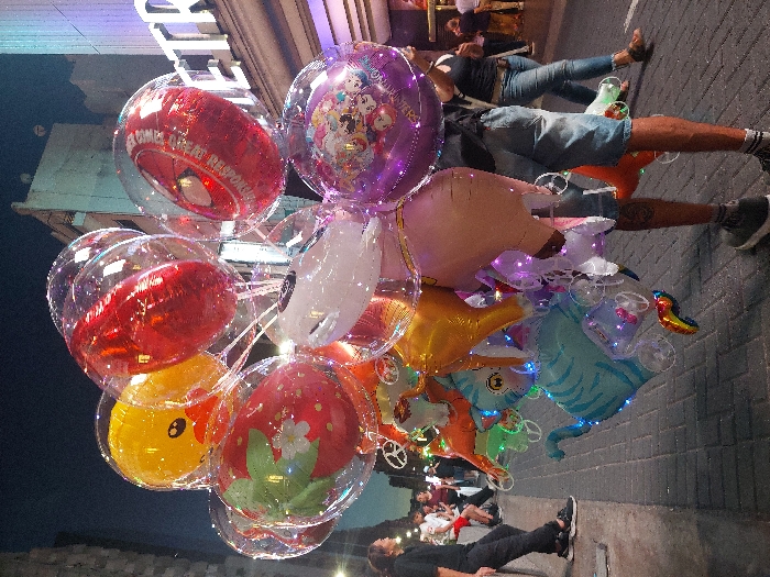 A cluster of vibrant, transparent balloons filled with various shapes and colors dominates the scene. Some balloons contain a red "Spider-Man" motif, a purple cartoon design, a yellow duck, and a strawberry with a white flower. Other balloons show animal figures, including a tiger. People are visible in the background, one in a black outfit leaning on a wall, and others seated at tables. The setting appears to be a street or public area, illuminated by streetlights.