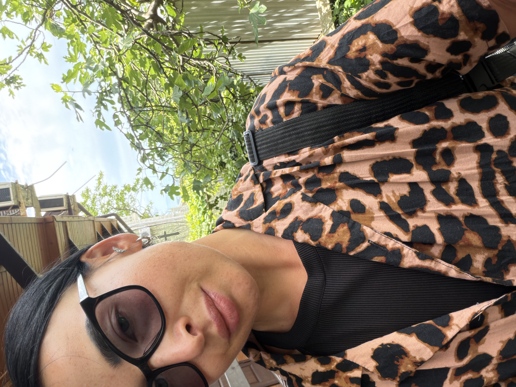 A medium-shot, selfie-style photograph captures a woman outdoors, likely in Sochi, Russia, given the lush green foliage and hints of a wooden building structure in the background. The woman, with dark hair pulled back and wearing large black-rimmed glasses, is looking towards the camera. She has on a black ribbed top under an open leopard-print shirt. A black strap, possibly from a bag, is visible across her shoulder. Her left ear is adorned with multiple stud earrings and a hoop earring with a cross charm. The background features green leaves from a tree and a corrugated metal fence, suggesting a natural or semi-urban setting. The lighting indicates it is daytime.