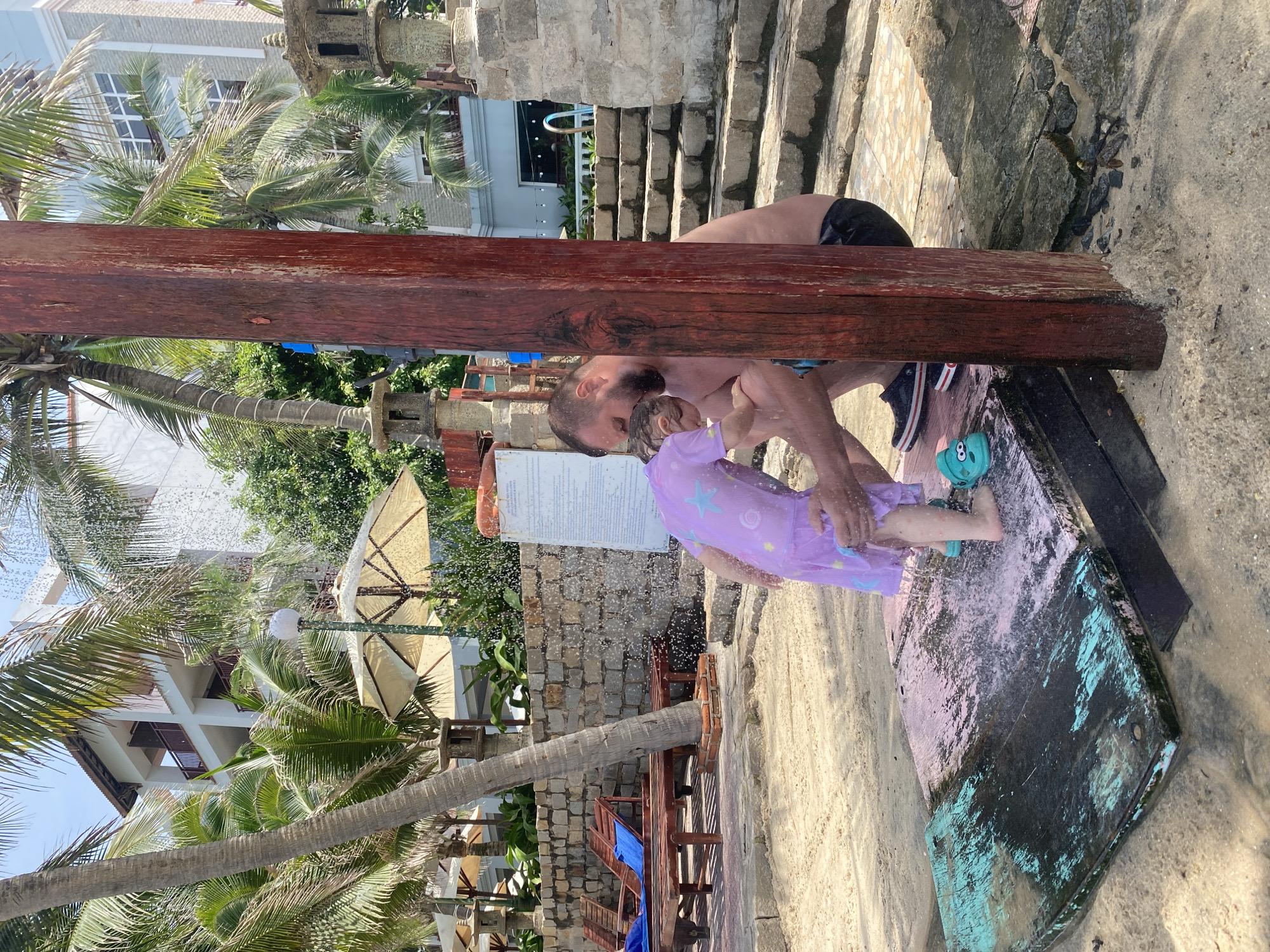 A man and a young child are being showered with water outdoors on a sandy beach. The man, with a beard, is crouching and appears to be washing the child, who is wearing a purple dress with a star pattern. They are standing on a wooden platform that is wet and appears to be partially submerged in water. The shower water is spraying onto them and the platform. In the background, palm trees, a stone wall, lounge chairs, a building, and stone lanterns are visible. The setting suggests a resort or public beach area in Phu Thuy Ward, Vietnam.