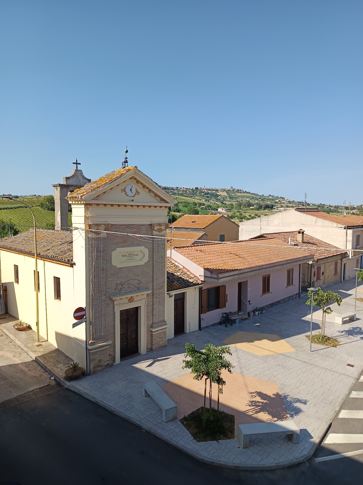 The image captures a sunny daytime scene in Roseto degli Abruzzi, Italy. The primary subject is a small church, likely a parish church, with a clock tower and a cross on top. The church's facade is adorned with a plaque. Adjacent to the church is a row of residential buildings with terracotta-tiled roofs. The street in front of the buildings is paved with gray tiles, featuring geometric inlays of light brown material around the bases of young trees. Benches are also positioned along the street. A red and white "no entry" sign is visible near the church. In the background, a hilly landscape is topped with a cluster of buildings, likely another part of the town. The sky is clear and blue.