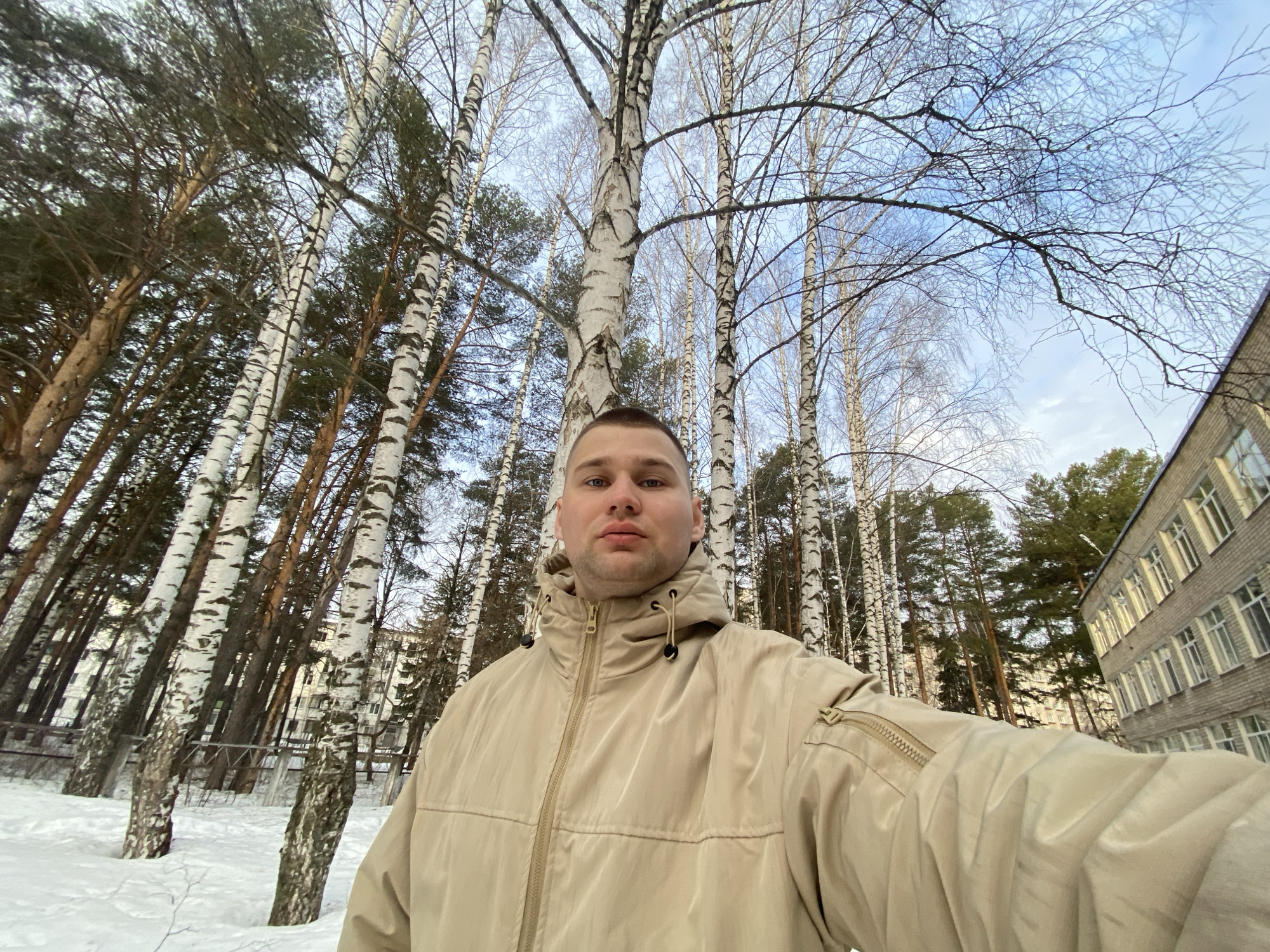 A male individual is centrally positioned, taking a wide-angle selfie outdoors in Chaikovsky, Russia. The subject has short, light-colored hair and light-colored eyes. He is wearing a light tan-colored hooded winter jacket with a visible central zipper and a zippered pocket on the left sleeve. The ground is uniformly covered by white snow. The background features a dense stand of trees, primarily birch trees with white and black bark and bare branches, intermixed with dark green conifer trees. To the far right, a multi-story building constructed of light-colored brick, featuring multiple rectangular windows with white frames, is visible. A faint, multi-story structure is also discernible in the distant background, partially obscured by trees. The sky above is light blue with scattered white clouds. A low, grey-colored metal fence runs horizontally through the mid-ground among the trees on the left side.