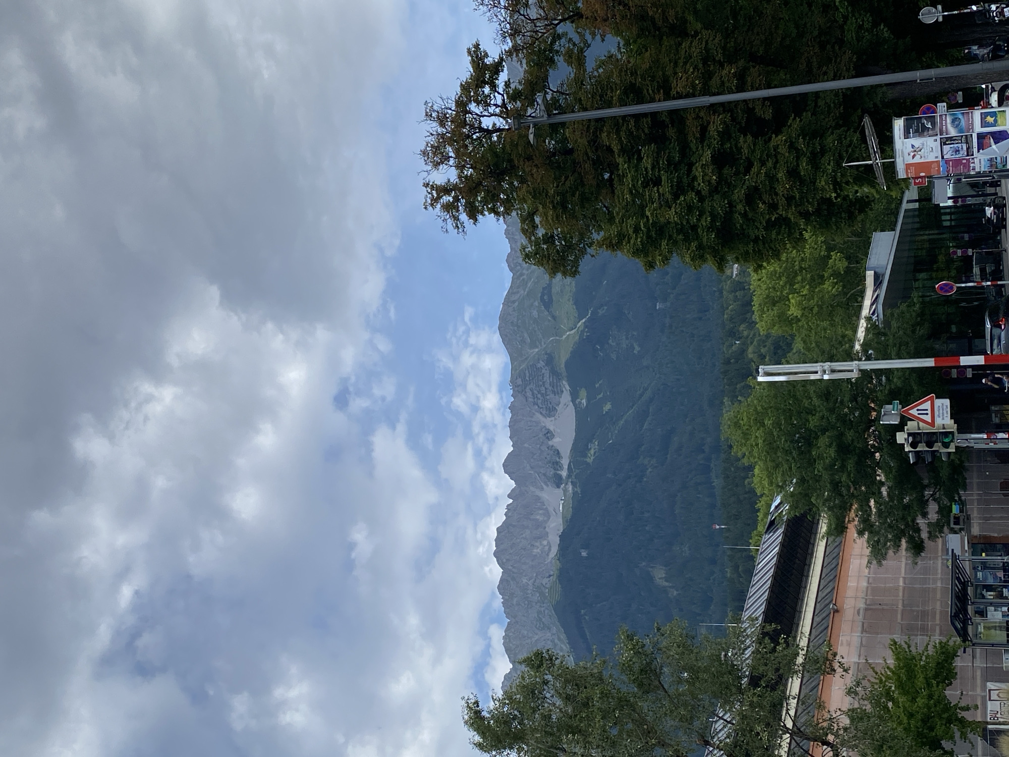 The image shows a view of the Nordkette mountain range from Innsbruck, Austria, on a cloudy day. In the foreground, there are trees, a street with traffic lights, a bus stop, and buildings. The Nordkette mountains are visible in the background, with their distinctive jagged peaks. The sky is filled with clouds, ranging from light and wispy to dark and heavy. The overall impression is one of a natural landscape meeting an urban environment.