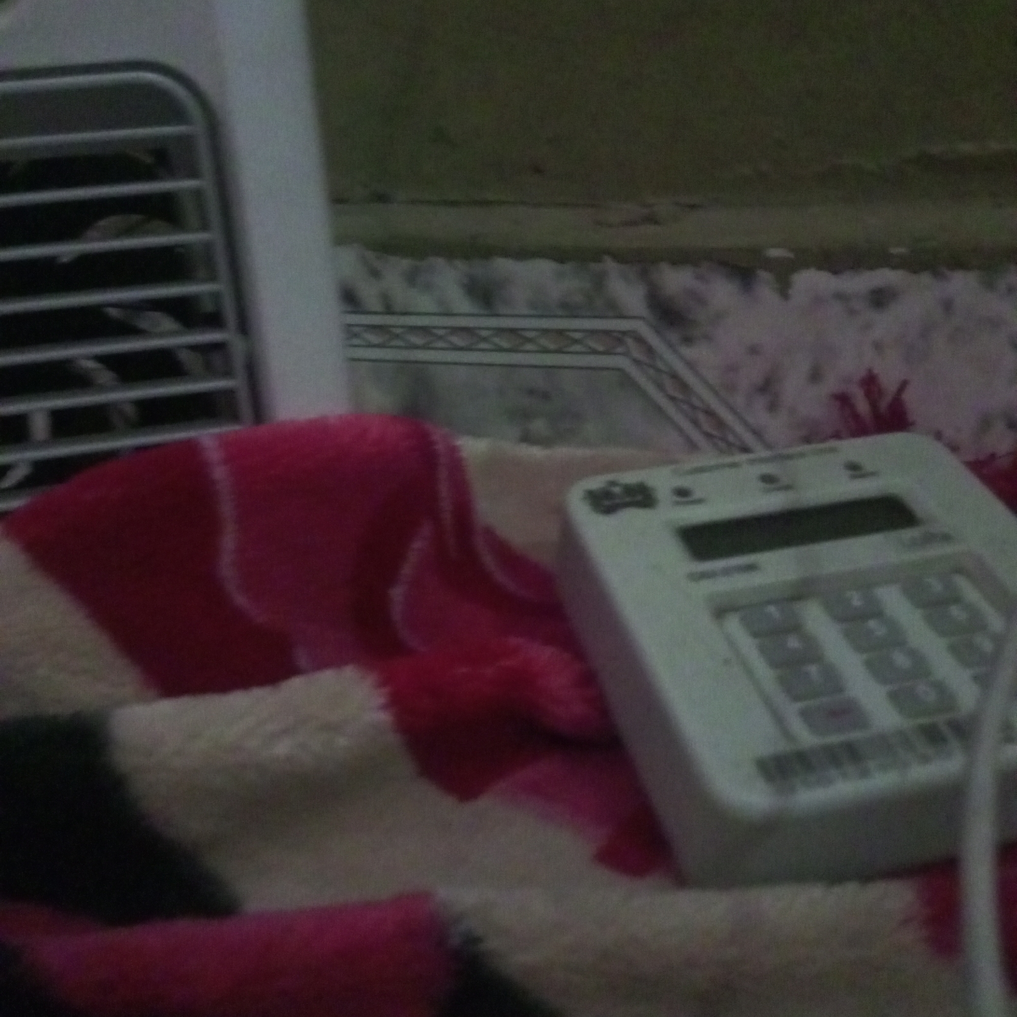 A close-up shot of a white keypad device with a dark screen sits on a pink and cream striped blanket. The keypad has numbers arranged in a grid, similar to a phone, and a small dark rectangular screen above them. The background is out of focus, but it appears to be a patterned carpet and a wall with some ventilation. The lighting is dim, suggesting it might be indoors or at night. The image is grainy and has low resolution. The location is Howayi, Nigeria.