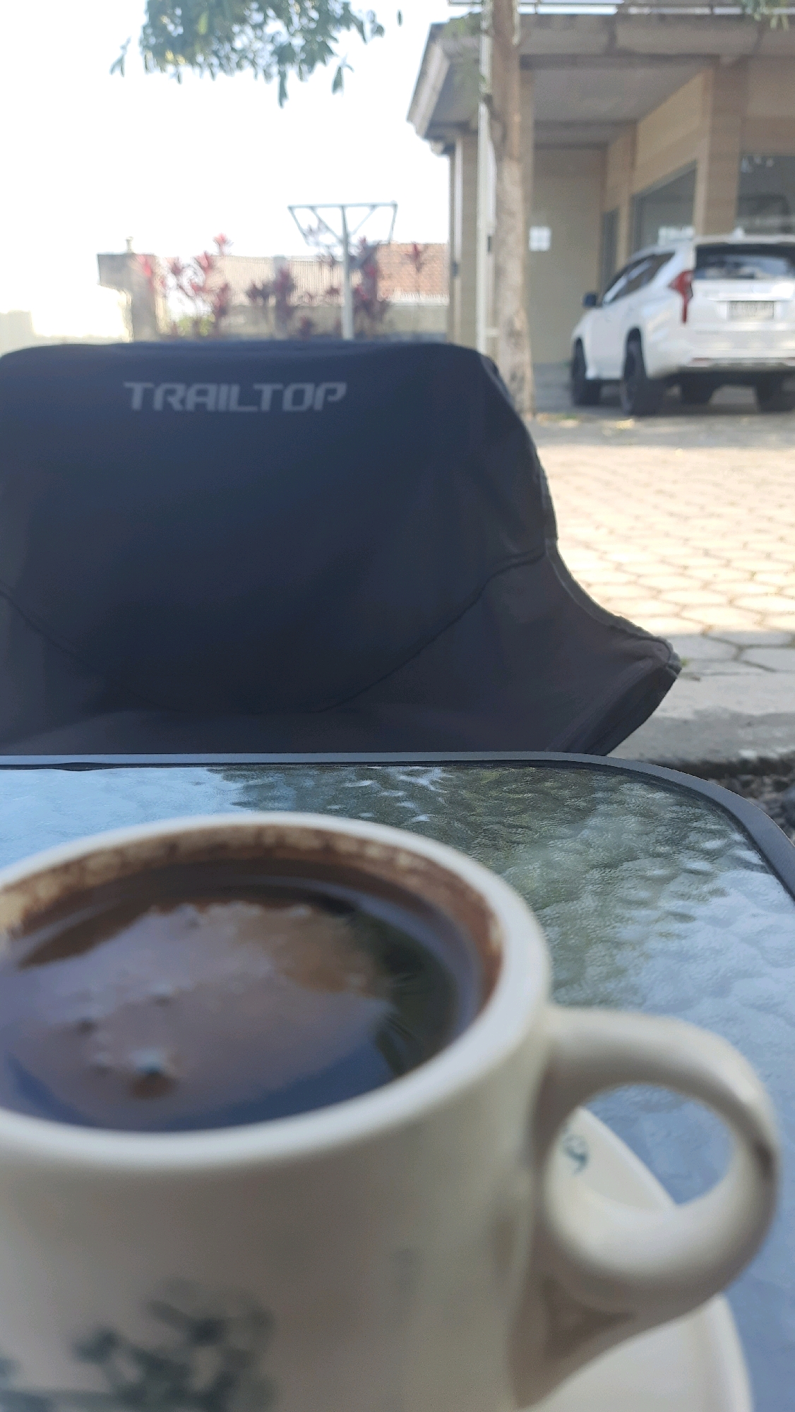 In the foreground of the image, a white ceramic cup filled with dark coffee sits on a glass-topped table. The table is outdoors, evidenced by the dappled sunlight reflecting on its surface. In the middle ground, a black bucket hat with the word "TRAILTOP" printed in white is placed on the table, partially obscuring the background. Behind the hat, a white SUV is parked in front of a building with a light-colored facade and windows. Red flowering plants are visible to the left of the building. The setting appears to be an outdoor area near a building, consistent with a residential or commercial establishment in Jajag, Indonesia. The overall lighting suggests it is daytime.
