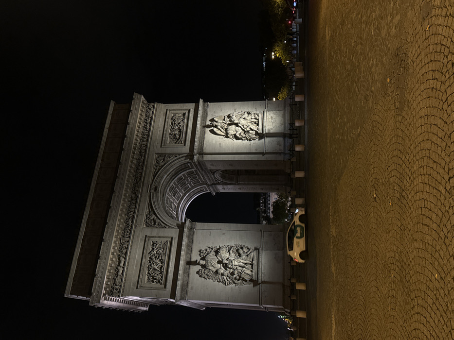 This is a nocturnal, wide-angle forensic photograph of the Arc de Triomphe, located in Place Charles de Gaulle, Paris, France. The massive neoclassical triumphal arch is centrally positioned and brightly illuminated by unseen floodlights, highlighting its stone construction, intricate sculptural reliefs on its facades (including "La Marseillaise" on the left and "The Triumph of 1810" on the right), and the coffered ceiling within its main archway. The sky above is completely dark and devoid of light sources or features.
In the foreground, a light-colored compact car, likely white or light grey, with a prominent green and white "Bolt" ride-sharing logo on its side, is captured in motion, exhibiting slight blur as it traverses from left to right across the frame. Its headlights are active, casting a beam forward. The ground beneath the car and extending into the immediate foreground is paved with dark, circular cobblestones, illuminated by ambient light sources to a warm, brownish-orange hue.
A line of light-colored, cylindrical bollards separates the cobblestone foreground from the base of the monument. Beyond the archway and to the right, dark trees and faint distant city lights are visible. The scene is devoid of any discernible individuals or other vehicles besides the aforementioned car.