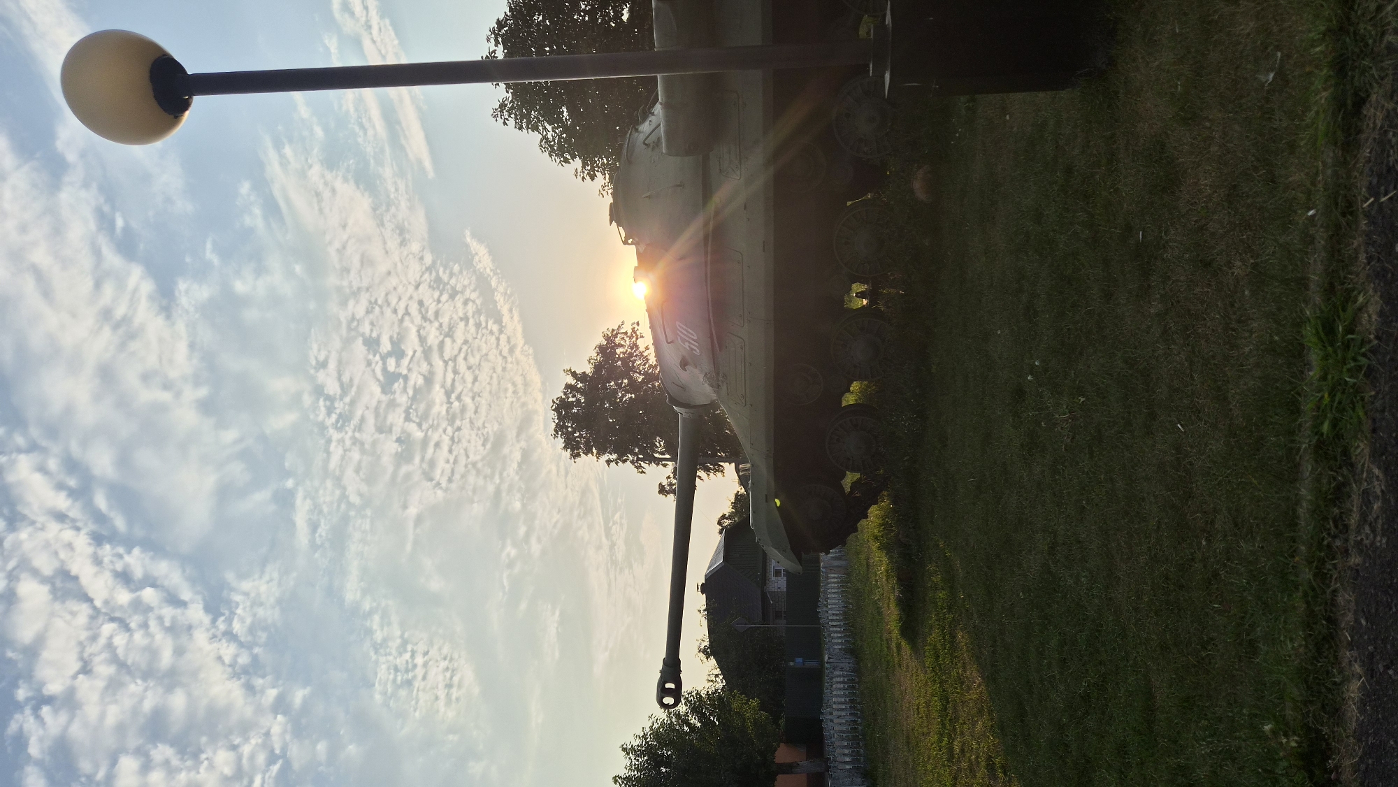 The image shows a military tank, identified as an IS-3, on display. The tank is positioned on a grassy area with a small white fence and some trees in the background. Residential buildings are visible behind the fence, suggesting a semi-urban or village setting. The sun is setting, casting a warm glow and creating lens flare as it peeks over the tank's turret. A lamppost is present on the right side of the frame, and the sky is filled with scattered clouds. The overall scene is peaceful, capturing a moment at dusk with a historical artifact as the focal point. The location is the village of Parichi, in the Parichi rural council of the Svetlogorsk District, Gomel Region, Belarus.
