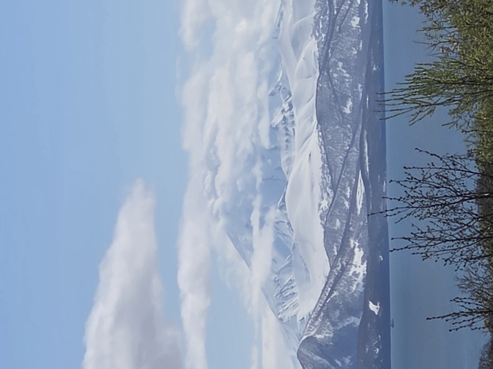 A landscape view is shown with a body of water, a snow-capped mountain range partially obscured by clouds, and some trees. The sky is blue with some clouds. The location is unknown, with the city also unknown.