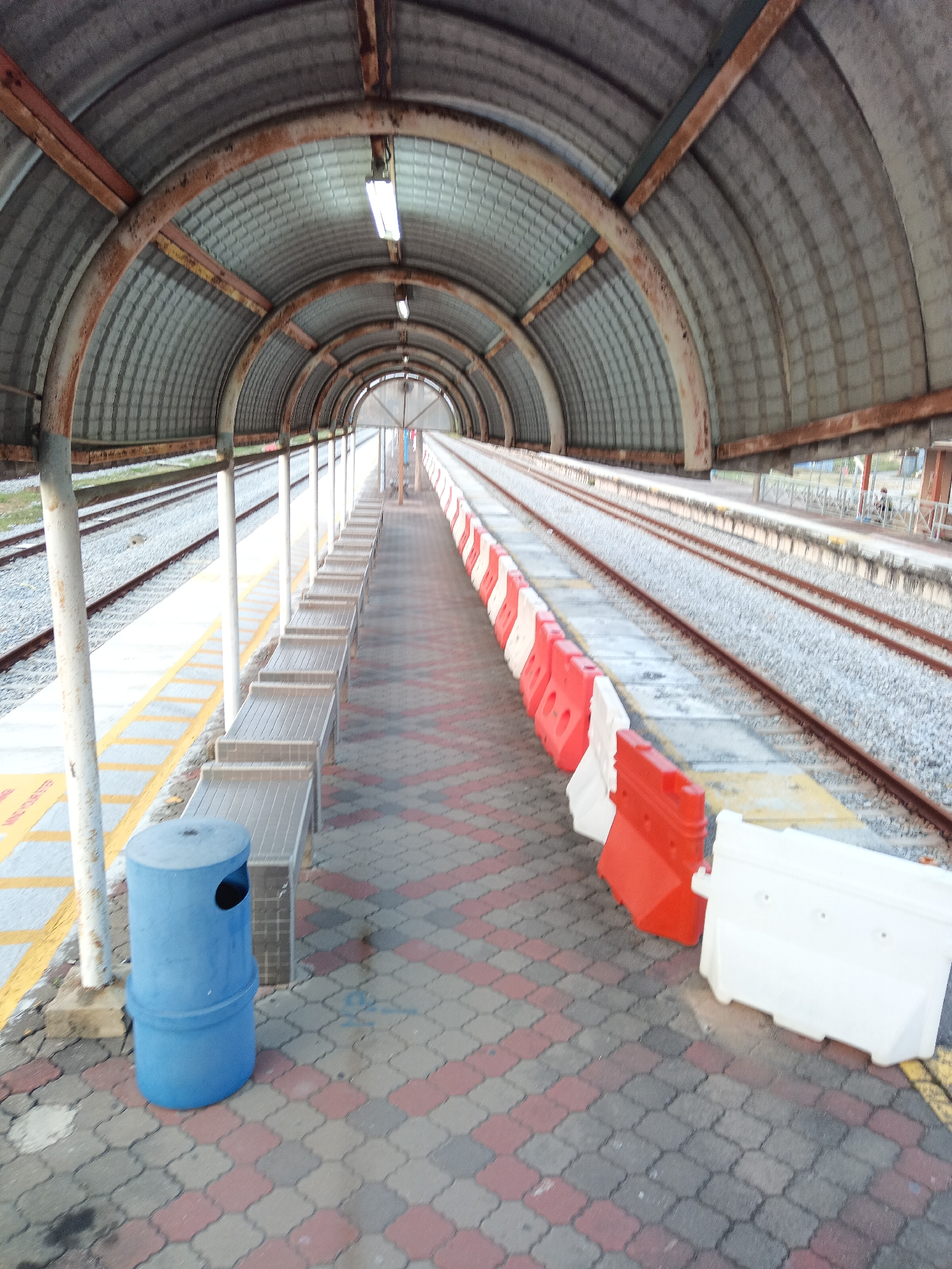 This is a wide-angle, eye-level shot of a covered outdoor walkway at a train station in Klang City, Malaysia. The walkway is covered by a corrugated metal roof supported by arched, rusty metal beams. Fluorescent lights are visible along the ceiling of the walkway. On the left side of the walkway, there are white vertical poles supporting the roof and several rows of gray benches. Adjacent to the benches is a yellow safety line on the platform, which is paved with patterned interlocking bricks in red and gray. A blue cylindrical trash can with a black lid is situated on the brick-paved area.
On the right side of the walkway, railway tracks are visible with ballast. The platform edge along the tracks is marked by a barrier composed of alternating red and white plastic construction barriers and white plastic Jersey barriers. The scene appears to be an active train station, with ongoing track maintenance suggested by the presence of the barriers. The overall atmosphere is utilitarian and functional, typical of public transportation infrastructure. There are no people clearly visible in the immediate foreground or middle ground of the image, but a distant figure can be observed in the background on the right side of the frame, near a building. The lighting suggests it is daytime, with diffused light entering from the open end of the walkway.