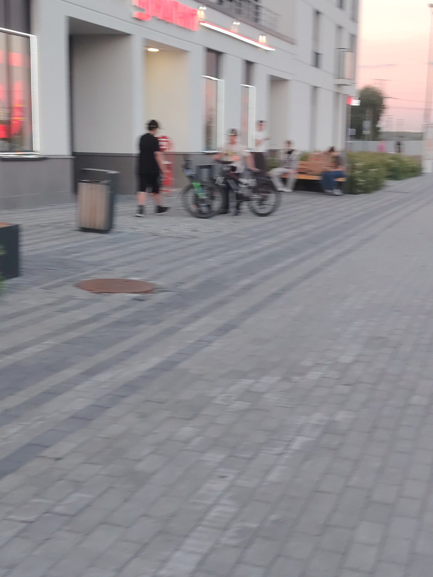 The image depicts a blurry outdoor scene in what appears to be an urban or suburban environment. In the foreground, a paved walkway with a distinctive brick pattern leads towards a modern building. To the left, a portion of the building's facade is visible, featuring large windows and what seems to be the edge of a store or commercial establishment with red lettering on its exterior. A trash receptacle with a wooden accent stands near the walkway.
Several individuals are present in the mid-ground. On the left, a person in dark clothing and a hat walks away from the camera. Further back, near the building, a group of people are gathered. Some are seated on a long bench, while others stand or appear to be near a bicycle. One person is riding a bicycle. The overall lighting suggests it might be late afternoon or early evening, with a soft, warm glow in the sky, possibly indicating sunset. The atmosphere appears casual, with people engaged in everyday activities like walking, sitting, and cycling. No specific location cues are immediately discernible beyond the general architectural style of the building and the paving. The blurry nature of the image prevents detailed observation of faces or specific objects.