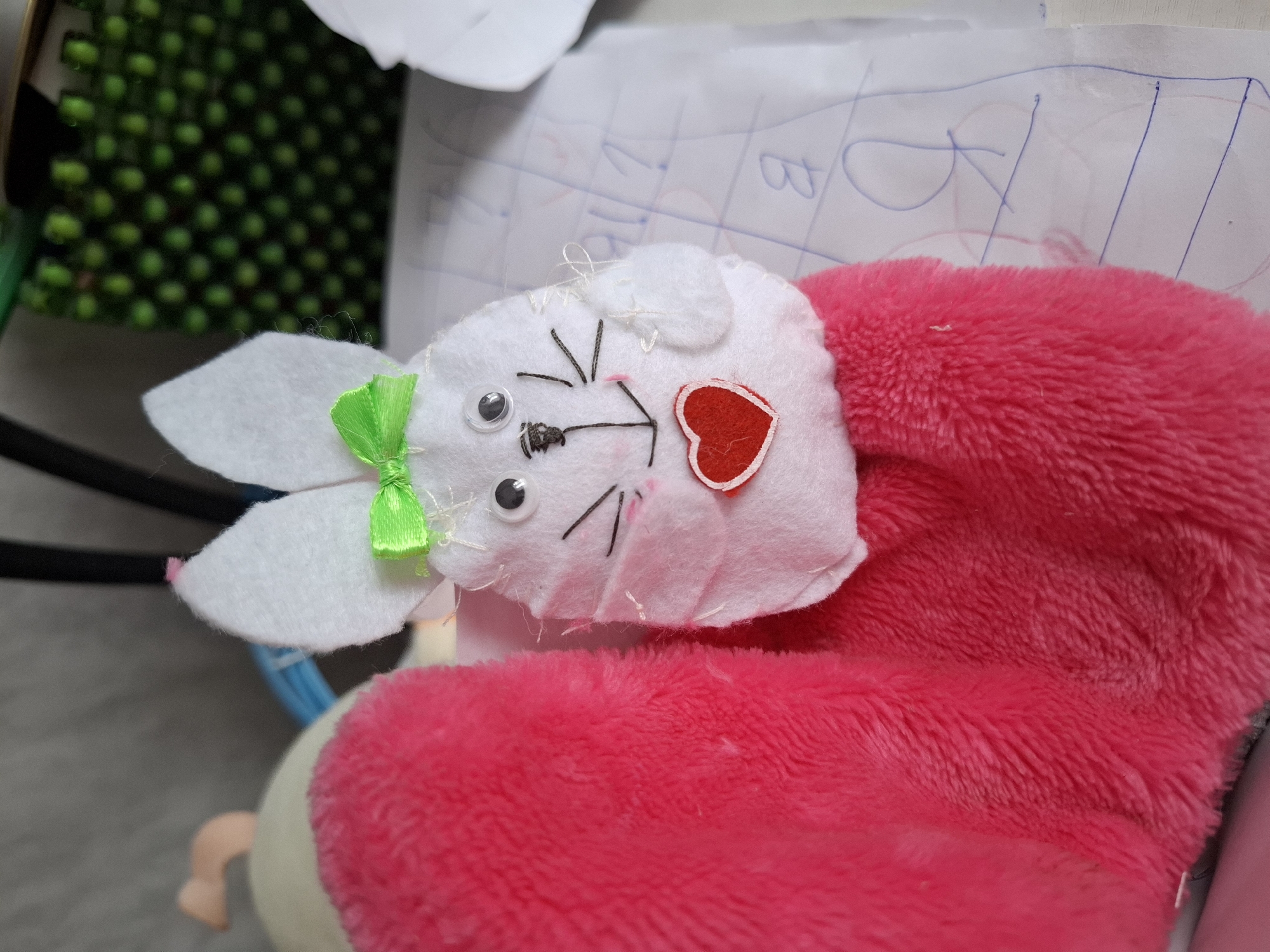 A close-up, top-down shot shows a handmade plush bunny toy, positioned to the left of the frame. The bunny has white felt ears, each adorned with a lime green ribbon tied into a bow. Its face is made of white felt with black embroidered whiskers and two googly eyes. A small, red felt heart is affixed to its right cheek, and its nose is a pink dot. The bunny's body is covered in fluffy, bright pink fabric. The background is blurred, but appears to be a surface with some handwriting and grid lines, possibly a piece of paper or a notebook. To the left of the bunny, a blurred section of a dark green, textured object is visible. The lighting suggests an indoor setting.