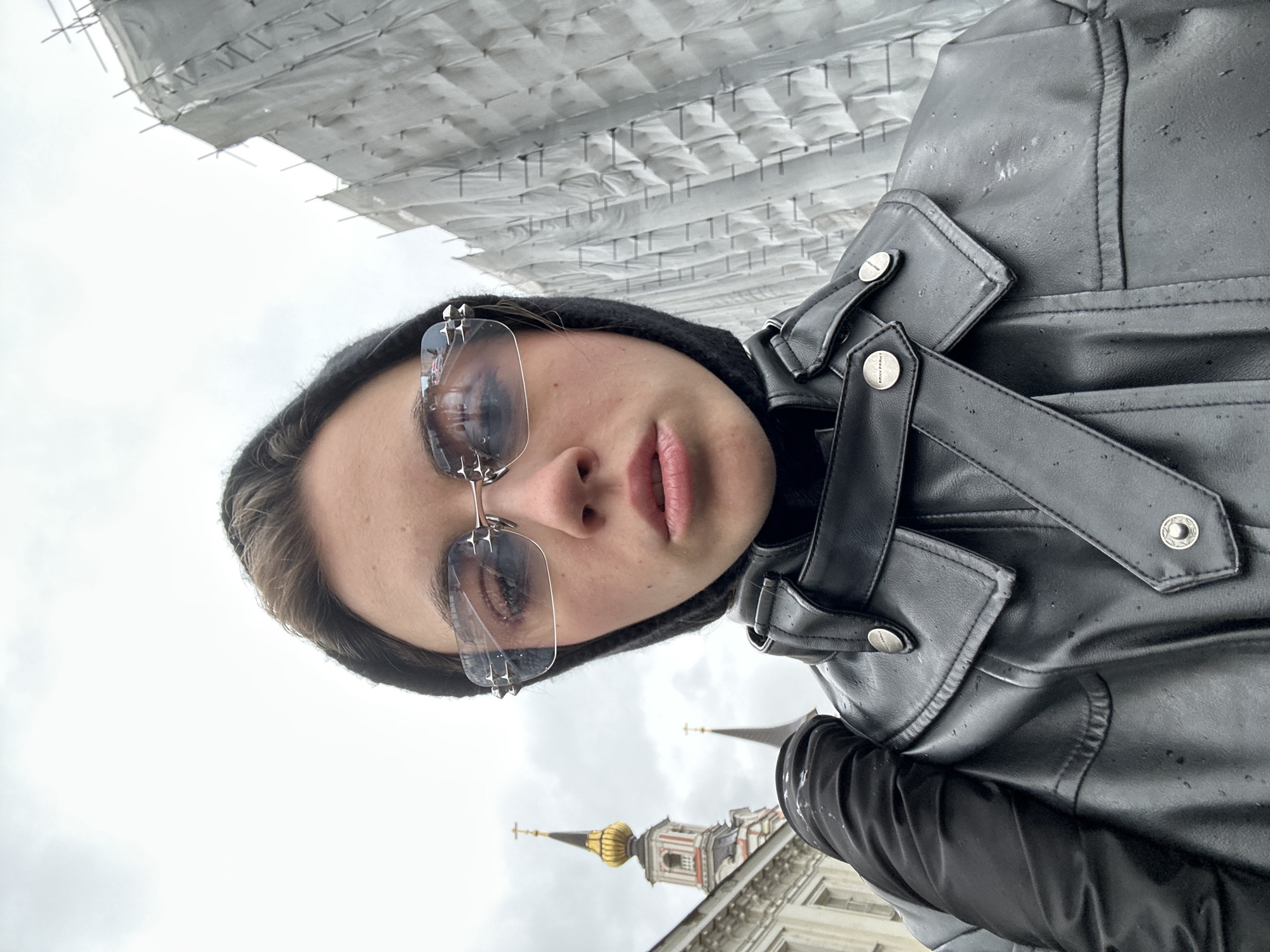 A low-angle, close-up, selfie-style portrait of a young woman wearing a black hooded garment, futuristic-looking, angular sunglasses with silver spike embellishments, and a black faux-leather jacket. She is looking directly at the camera with a neutral expression. Her face is centered, and her shoulders and upper chest are visible. The background features a cloudy, overcast sky. To the left of the woman, a portion of a domed, religious building with a golden dome and crosses can be seen. To the right, a large structure covered in white tarpaulin, possibly under construction or renovation, is visible. The scene suggests an outdoor urban setting, potentially in Moscow, Russia, given the architectural style of the building.