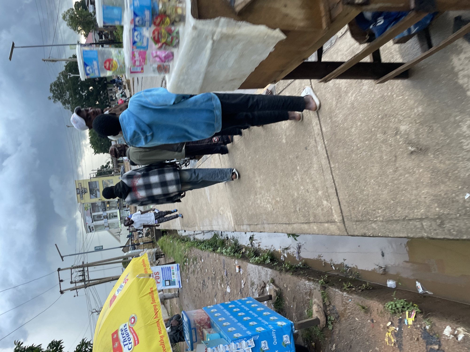 In Jos, Nigeria, a group of individuals walks along a sidewalk adjacent to a street. A yellow umbrella displaying the "Three Crowns" logo stands near the foreground, beside a vendor's stall displaying various items, including bottles of "Peak Milk." Further down the sidewalk, other people are seen walking, and a vendor appears to be attending to customers. The environment includes a drainage ditch filled with murky water and some debris, and buildings and electrical poles are visible in the background. The sky is overcast, suggesting a cloudy day.