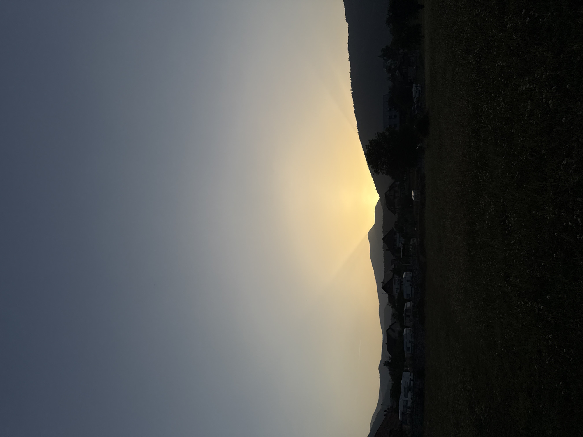 The image depicts a twilight scene in Plav, Montenegro. The sky transitions from a deep blue at the top to a soft yellow near the horizon, where the sun is setting behind a mountain range. Rays of light fan out from behind the mountains, illuminating a portion of the sky. Silhouetted against the illuminated sky are rolling hills and a village nestled at their base. Several houses with visible rooftops are clustered together. In the foreground, a dark, grassy field stretches across the frame, dotted with small, light-colored wildflowers. Row of caravans are parked in front of the village. The overall atmosphere is serene and tranquil, typical of the evening in a rural mountainous region.
