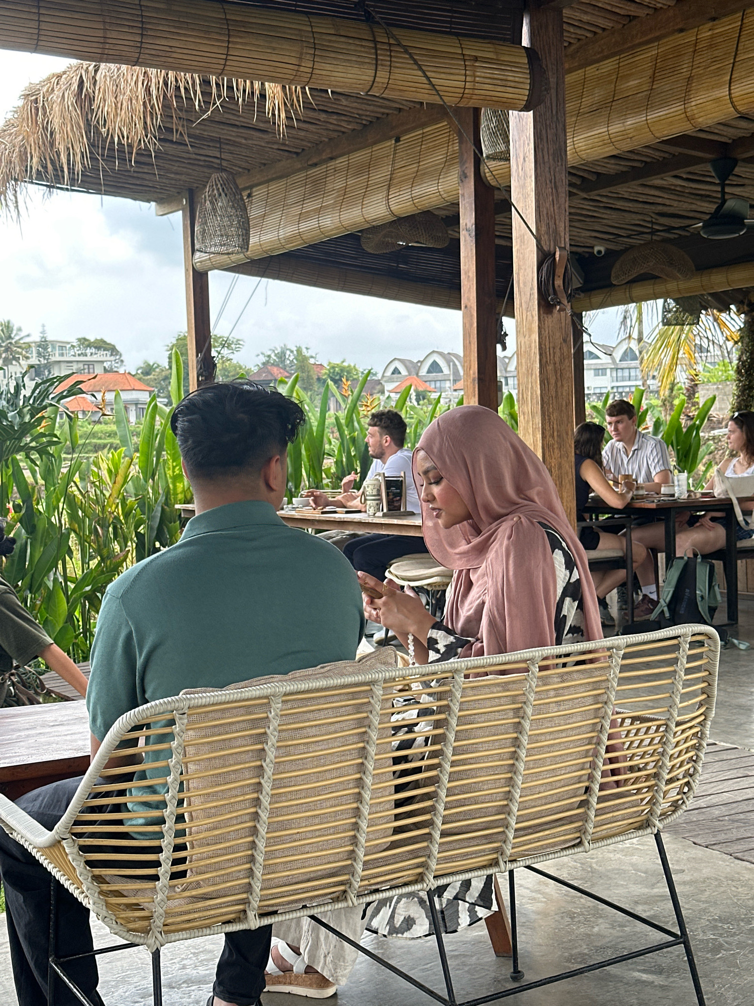 This is a medium shot of a couple sitting on a woven bench in an outdoor restaurant with a thatched roof and bamboo blinds. The couple is facing away from the camera. The man, on the left, is wearing a teal polo shirt and has dark hair. The woman, on the right, is wearing a pink hijab and a black and white patterned dress. She is looking down at her phone. In the background, there are other diners at tables, lush green vegetation, and buildings. The lighting is overcast.