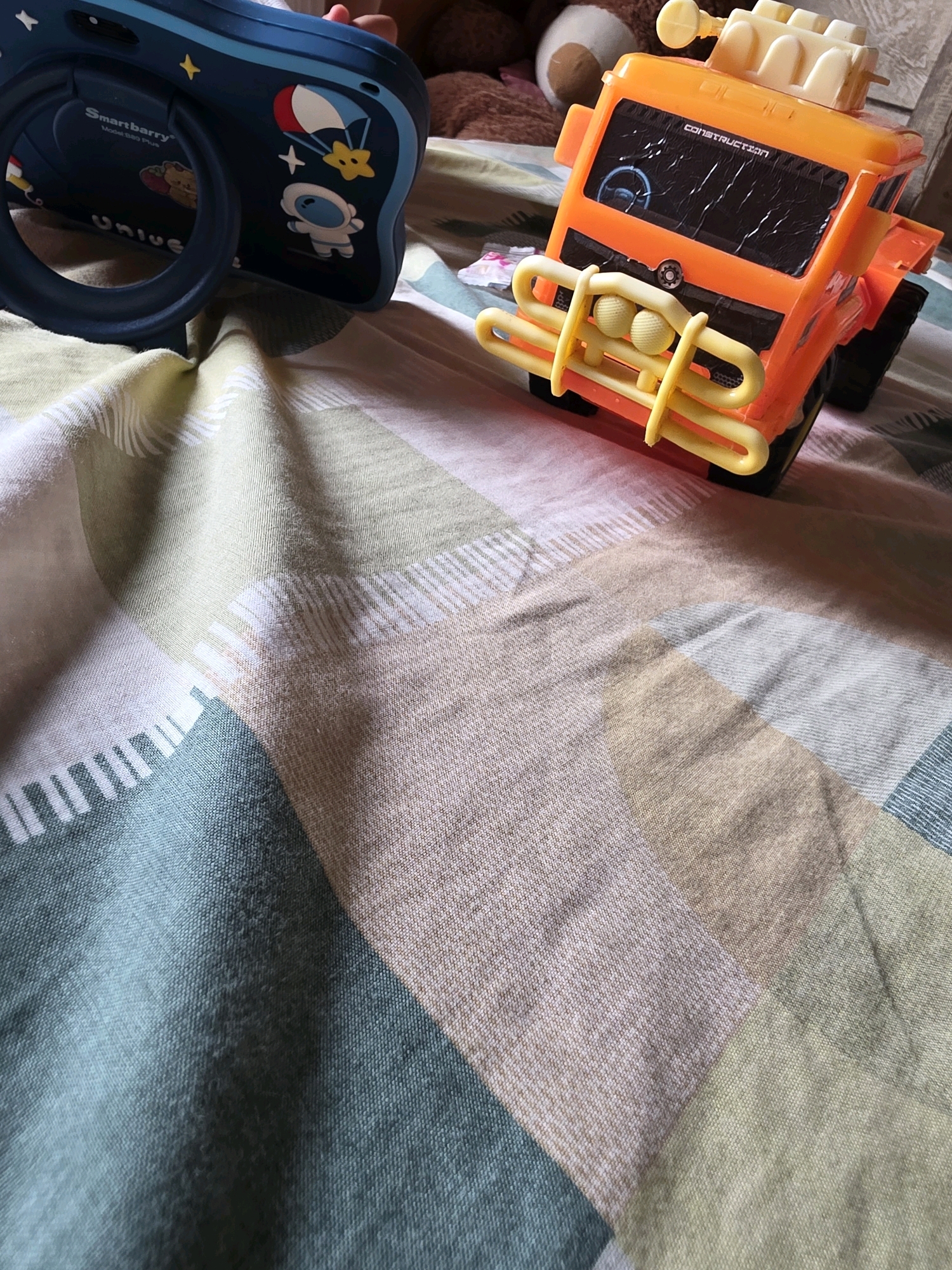 A close-up, high-angle shot shows two children's toys on a bed with a patterned comforter. On the left is a dark blue toy that appears to be a camera or similar device, decorated with space-themed stickers including an astronaut and a parachute. To the right is a bright orange toy construction truck with a yellow bumper. The comforter is a patchwork of beige, light green, and muted teal blocks with white stripes. The scene is indoors, likely in a child's room, but no specific location in Kota Tanjung Selor, Indonesia can be determined from the image.