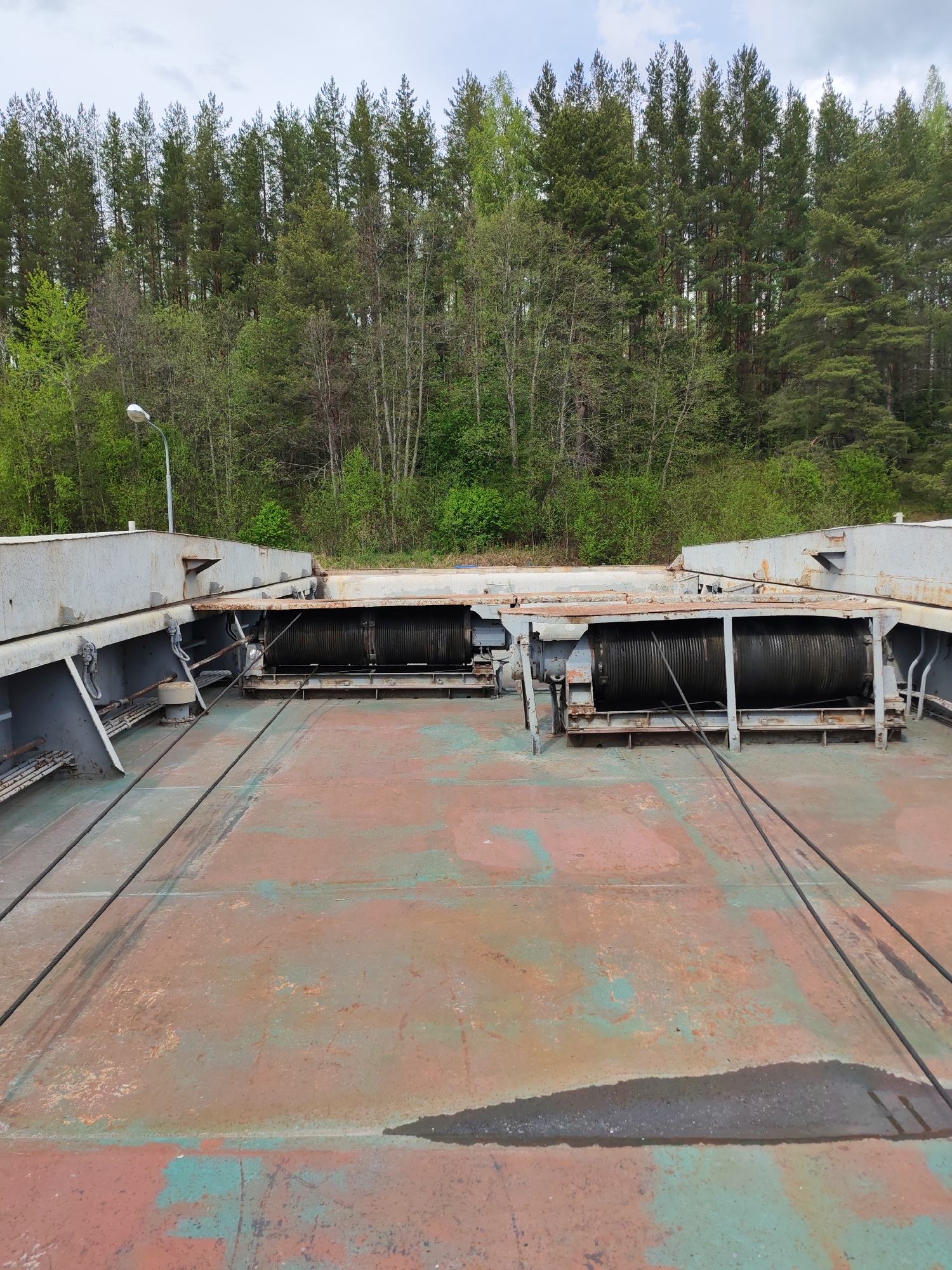 The image shows a section of what appears to be a dam or large industrial structure in Alekseevskoe, Russia. Two large, rusted metal winches with wound cables are visible in the center, anchored to a gray metal structure. The floor is a large expanse of rusted metal plating with patches of green and red paint. In the background, a dense forest of coniferous trees is seen, and a solitary street lamp extends from a structure on the left. The sky is overcast. No people are visible.