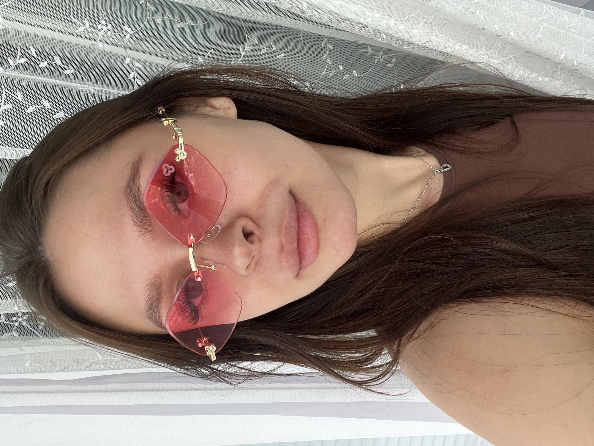 The media file is a close-up, vertically oriented selfie of a young woman. She has long, dark brown hair that is spread out around her. She is wearing large, pink, heart-shaped sunglasses with gold detailing. A silver necklace with a small pendant is visible on her neck. She is wearing a brown t-shirt or top. The background appears to be a white, sheer curtain with a floral pattern, likely indoors. The lighting suggests it is daytime, and the overall mood is relaxed and casual. There is no visible text in the image. The setting appears to be domestic, possibly a bedroom or living room in Kazan, Russia.