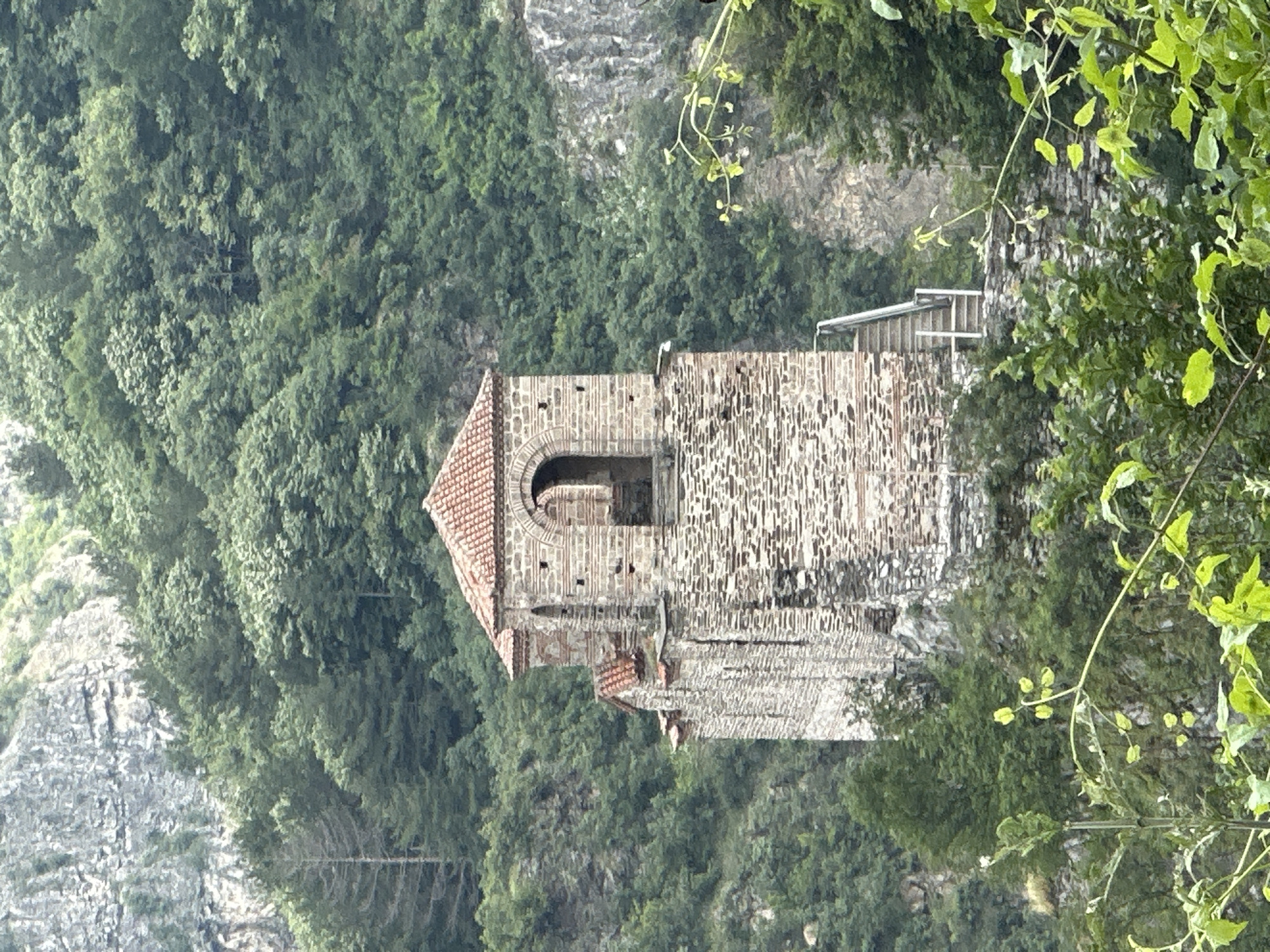 An ancient-looking stone tower or building is the central subject of this image, nestled within a lush, forested mountain landscape. The structure is built from rough, multi-toned stone blocks, displaying traditional masonry. It features a distinctive reddish-brown tiled roof and an arched opening, possibly a window or a doorway, on one side. A smaller, lower section of a wall or another part of the building, also constructed of stone, extends to the right.
The building is surrounded by dense green foliage, with numerous deciduous trees covering the steep hillsides that form the background. Rocky outcrops and natural cliffs are visible through the trees, particularly in the upper right portion of the scene, highlighting the rugged terrain. In the immediate foreground, blurred green leaves and branches partially obscure the view, suggesting the photo was taken from a vantage point surrounded by vegetation.
No specific activity or individuals are present, lending a tranquil and enduring atmosphere to the scene. The lighting is soft and even, characteristic of an overcast day or late afternoon, without harsh shadows. The combination of historical architecture and natural beauty points to a location like Asenovgrad, Bulgaria, which is known for its historic fortresses and monasteries integrated into mountainous settings. There is no visible text within the image.