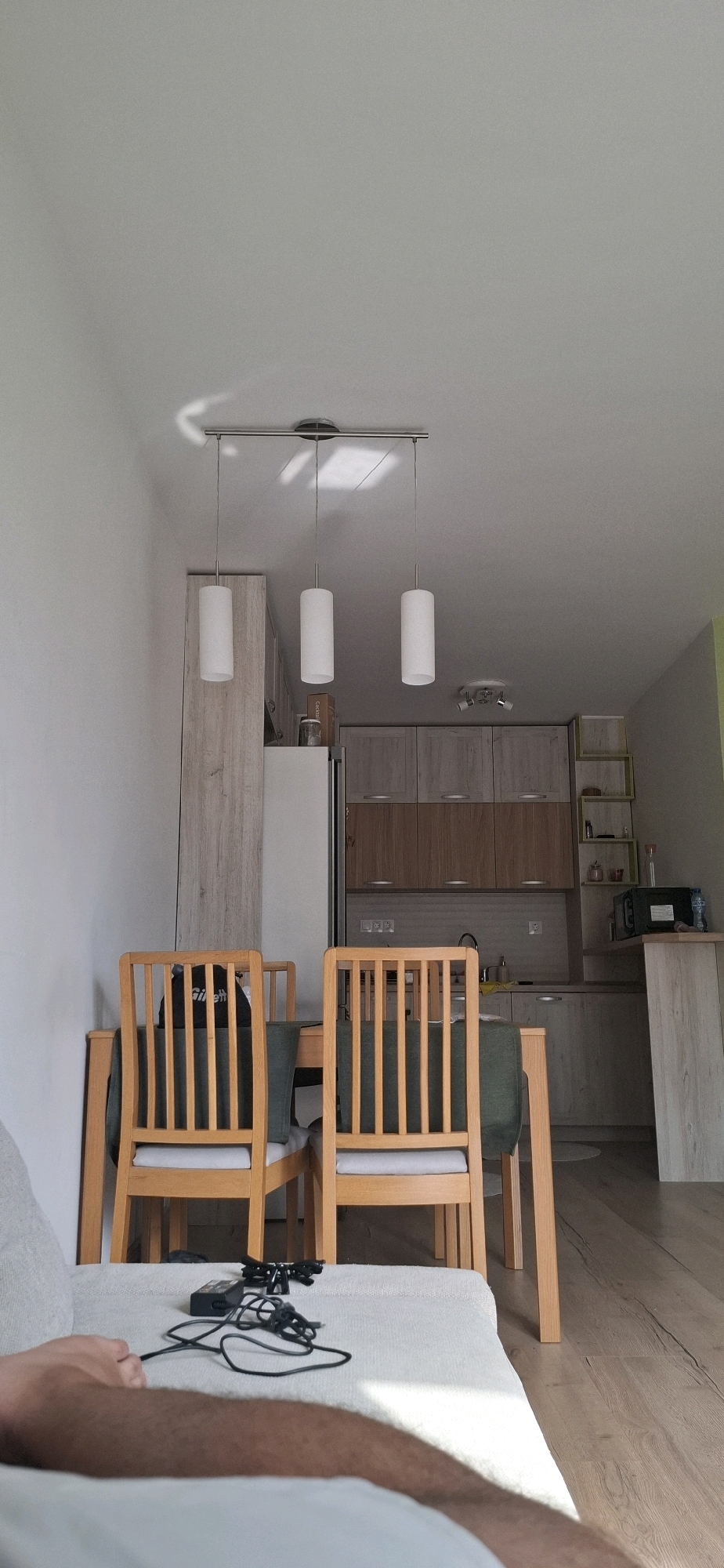 This image captures a domestic interior scene, likely a living or dining area adjacent to a kitchen, in a city apartment in Sofia, Bulgaria. The perspective is from a low vantage point, suggesting the photographer is seated or lying down on a couch or bed in the foreground.
The foreground is dominated by a portion of a person's leg and arm, covered by a light-colored fabric or blanket. In front of this, on a similar light-colored surface, are electronic cables and a black rectangular device, possibly a charger or remote control.
Further back, a wooden dining table with a dark green tablecloth is visible. Two light-colored wooden chairs with slatted backs and light grey seat cushions are positioned at the table. One chair has a black bag with "Gillette" printed on it resting on its seat.
The background reveals a kitchen area. To the left, a tall, light wood-paneled cabinet or refrigerator stands against a white wall. In the center, kitchen cabinets with light wood-effect doors and darker brown lower cabinets are mounted above a countertop. A sink and faucet are visible on the countertop. To the right of the cabinets, a narrow, light wood shelving unit with open shelves is present. On the countertop near the shelving unit, a black microwave oven and a water bottle are visible.
Above the dining table and kitchen area, three white cylindrical pendant lights hang from a metal track mounted on the ceiling. The ceiling is plain white. The walls are painted white. The flooring in the dining and kitchen area appears to be light wood laminate. The overall lighting suggests a naturally lit interior, with some shadows cast from the pendant lights. The scene depicts a functional, modern living space.
