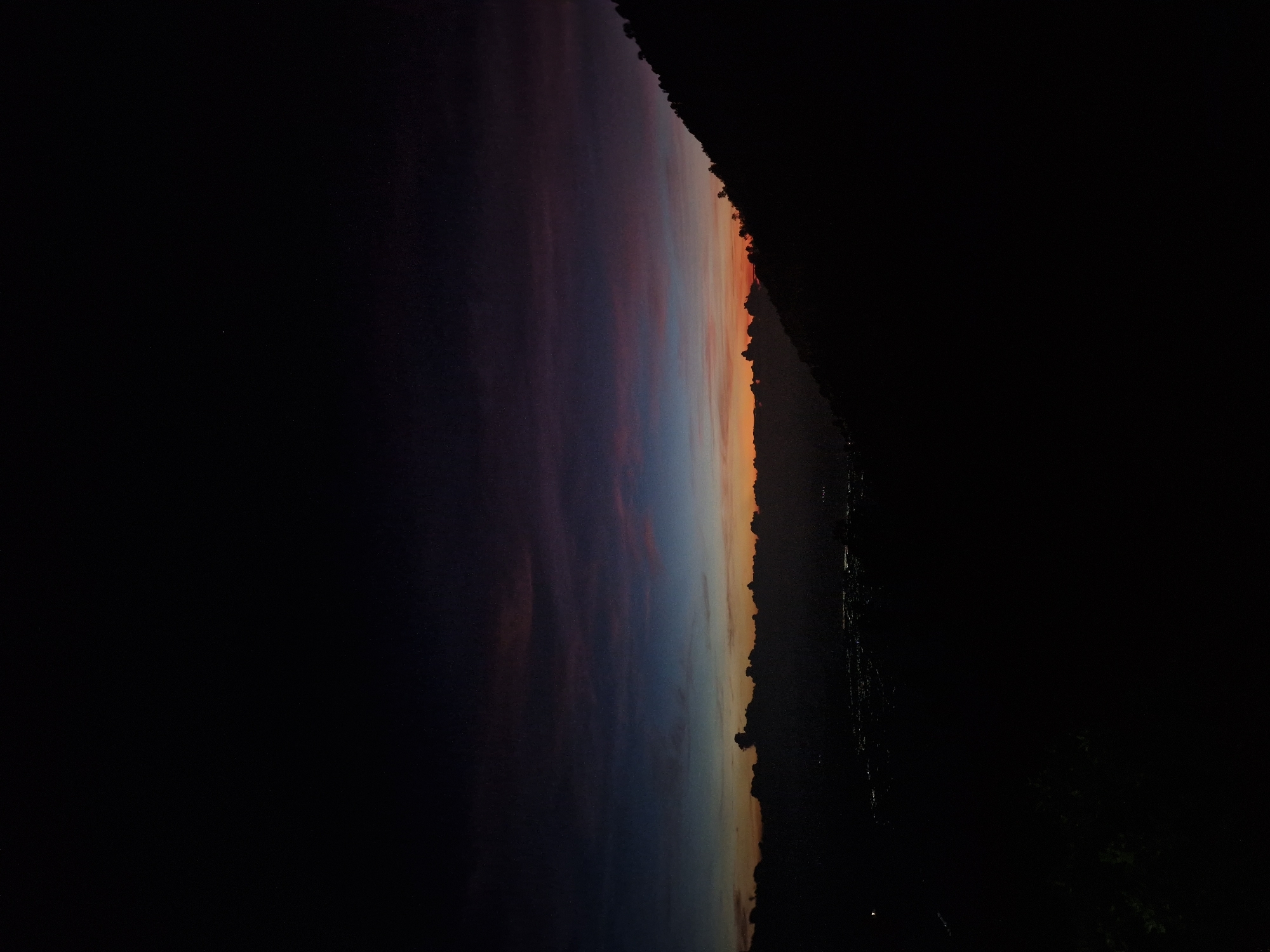 The image shows a landscape at dusk in Unknown, Indonesia. The sky transitions from orange on the right edge to blue and purple on the left, with clouds visible. A dark silhouette of trees or a hillside is on the right. The ground below appears black. Tiny lights are scattered in the silhouette.
