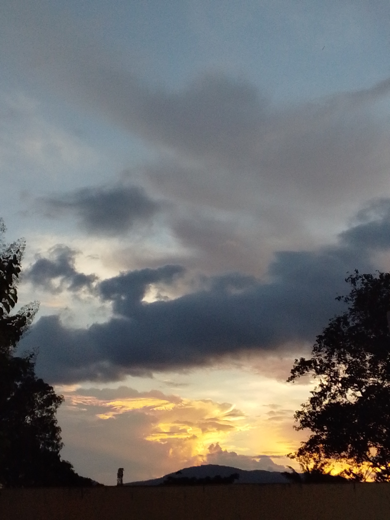 The image captures a serene sunset scene in Serti, Nigeria. The sky is adorned with dramatic clouds, ranging from soft grays to deep blues, illuminated by the warm hues of the setting sun. The lower part of the sky features a vibrant display of oranges and yellows, suggesting the sun is on the horizon. Silhouetted against this colorful backdrop are trees framing the scene on the left and right, their dark forms creating a stark contrast with the sky. In the middle ground, a solitary figure stands on a ridge, a small, dark silhouette against the bright sunset. Further in the distance, the gentle contours of hills are visible, also silhouetted. The foreground consists of a dark, horizontal band, likely representing the ground or a building's roof, with a textured, possibly dry, surface. The overall impression is one of natural beauty and tranquility as day transitions into night in Serti.