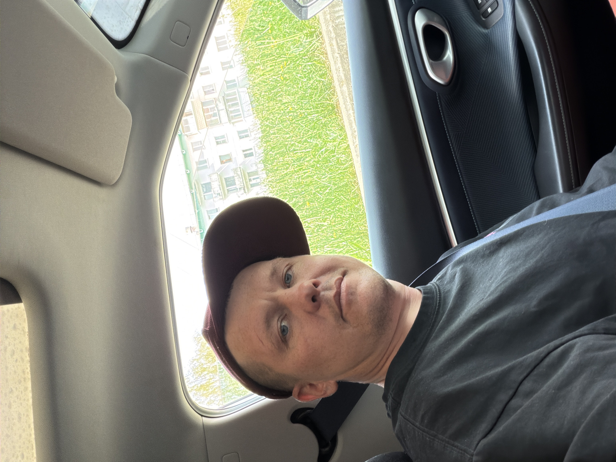 A man wearing a dark-colored t-shirt and a baseball cap is sitting in the backseat of a car. He is wearing a seatbelt. The car is moving, as indicated by the blurred background visible through the window, which shows a building and grass. The specific location of the photo is not verifiable as Chaikovsky, Russia, from the image alone. The interior of the car is visible, including a door panel. The man is looking at the camera.