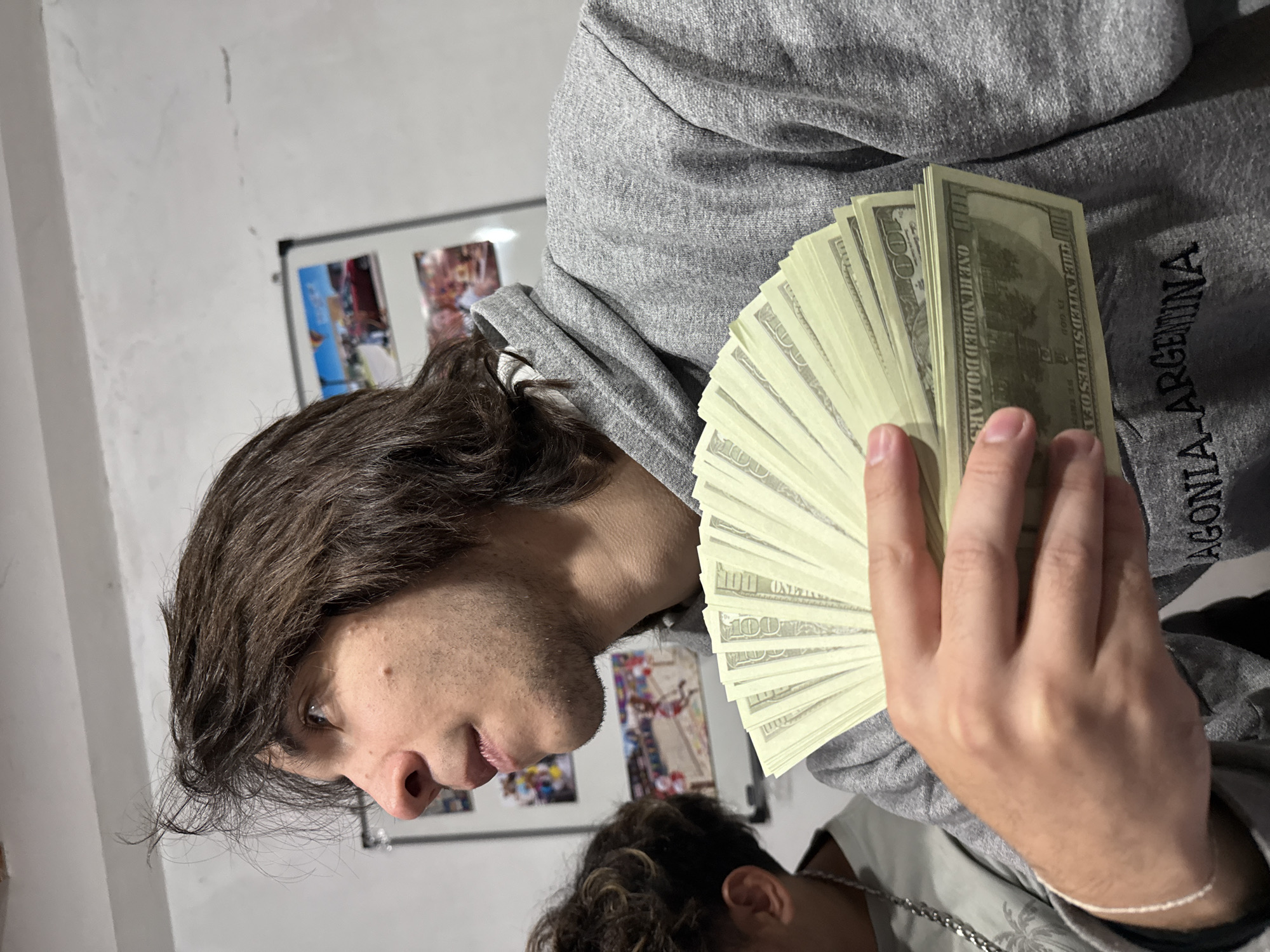 A young man with dark hair is holding a large fan of US hundred dollar bills. He is wearing a grey hoodie with the word "Patagonia - Argentina" printed on it. Another person is partially visible at the bottom of the frame, wearing a light colored shirt and a silver chain. A white wall is behind the man, with a framed photograph or poster visible on the wall. The location, San Fernando del Valle de Catamarca, Argentina, is indicated by the writing on the hoodie.