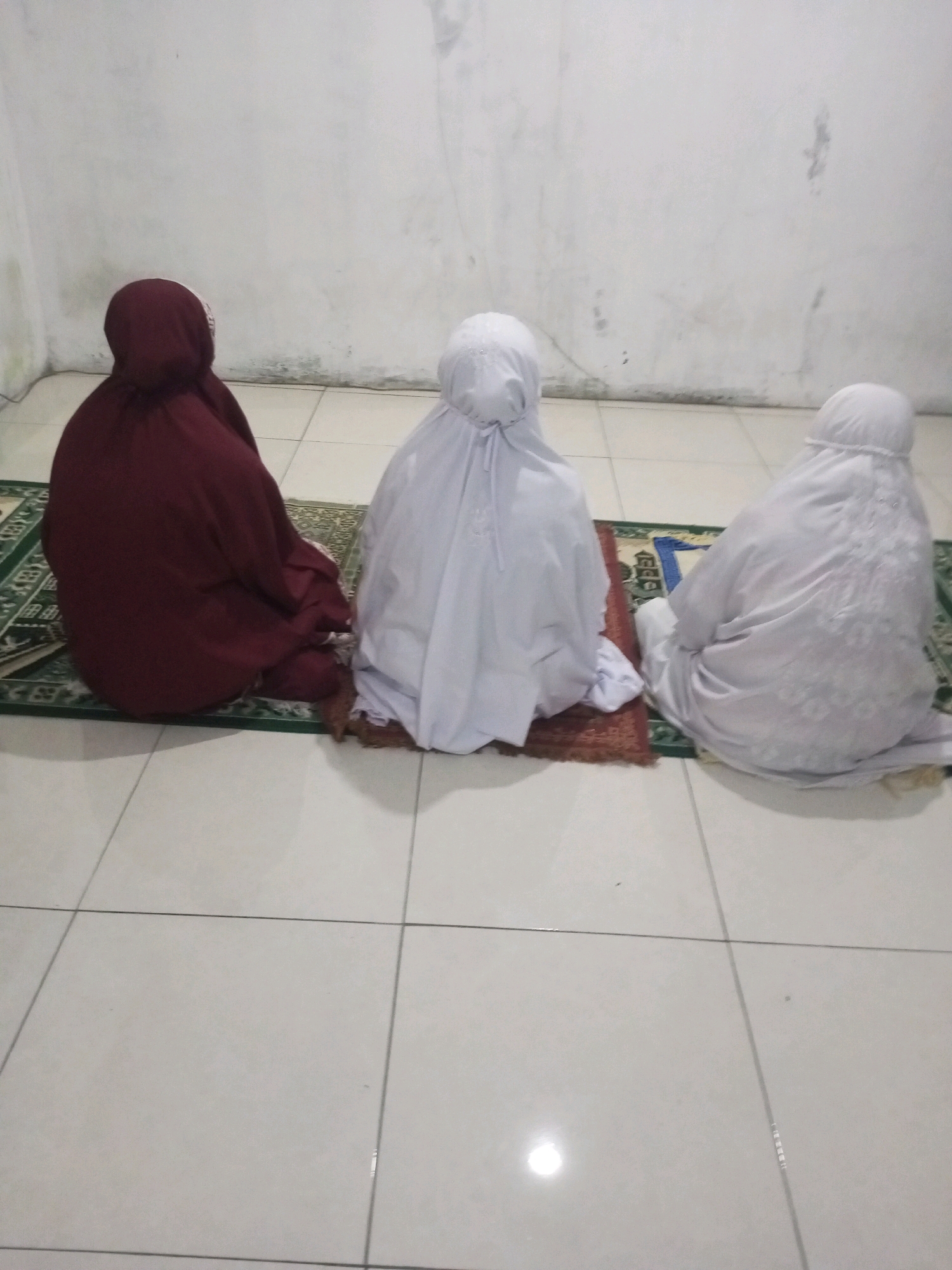 Three individuals, viewed from behind, are seated on prayer mats on a tiled floor. The individual on the left wears a maroon hijab. The two individuals to their right wear white prayer garments. The wall behind them is white and has visible scuffs and marks. The scene appears to be indoors.