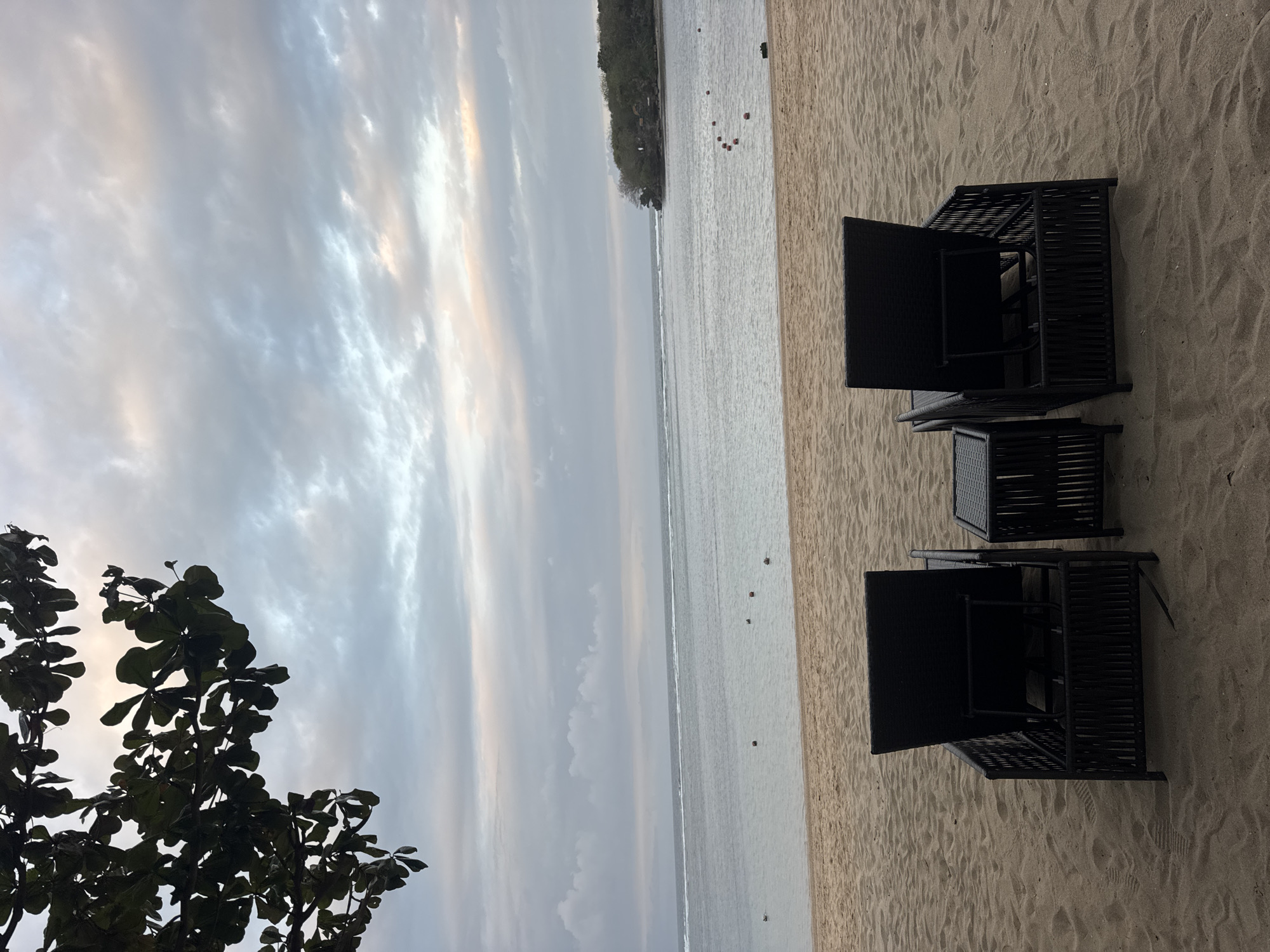The image depicts a serene beach scene in Kuta Selatan, Indonesia, likely at dawn or dusk due to the soft, diffused lighting. In the foreground, two dark-colored, modern lounge chairs made of woven material sit on golden sand. Between them, a small, matching dark-colored side table. The chairs are positioned to face the calm ocean. The sand shows subtle footprints, indicating previous activity.
The middle ground is dominated by the tranquil sea, its surface reflecting the muted colors of the sky. A thin, white line of surf marks the horizon where the sea meets the sky. Several small, reddish-orange buoys are visible in the water, suggesting a designated swimming or boating area. In the distance, a lush green island or peninsula, covered with trees, juts out into the water on the right side of the frame.
The sky is a canvas of soft, muted colors, with layers of clouds in shades of grey, white, and hints of pastel pink and yellow. The clouds appear textured and atmospheric, hinting at either an approaching or receding weather system. In the upper left corner, the dark silhouette of a tree branch with broad, green leaves hangs over the scene, providing a natural frame and a sense of natural beauty. The overall atmosphere is one of peace and quietude, inviting relaxation.