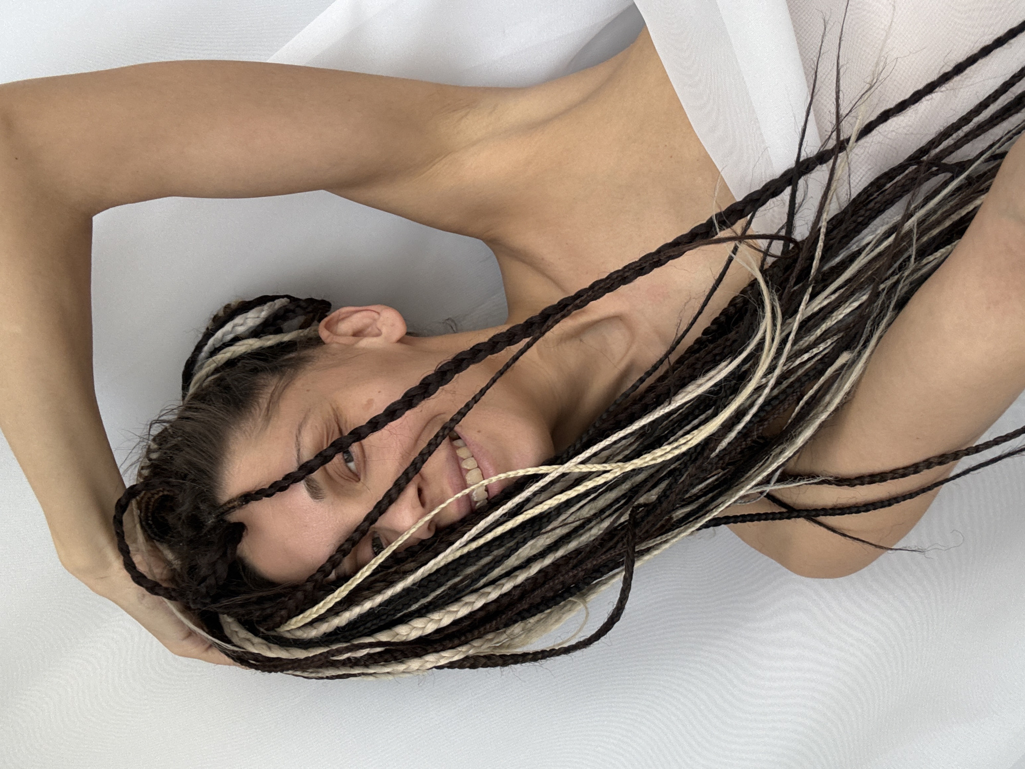 A woman with long, dark and light-colored braided hair lies on a white surface. Her head is tilted, and she is smiling, revealing her teeth. She has an arm raised on either side of her head. Part of a white cloth drapes over her. The photo appears to be taken from directly above the subject. There is no indication this image was taken in Rusanovo, Russia.