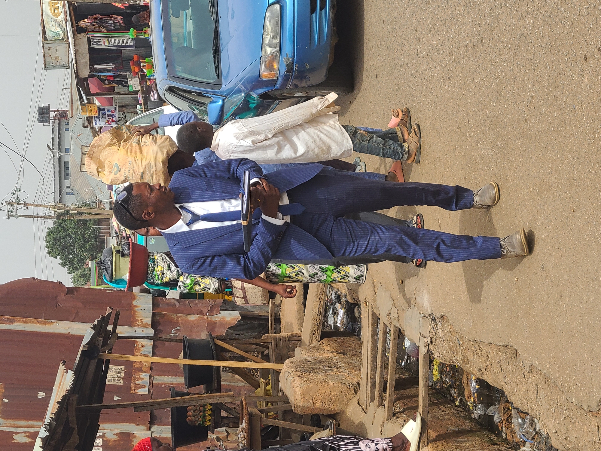 A man in a blue pinstripe suit and grey shoes stands in a street in Jos, Nigeria. He is holding a dark object. Behind him, two other men are carrying a large sack. One wears a white shirt. A blue car is parked nearby. The street is lined with buildings made of corrugated metal and wood, including what appear to be makeshift shops. A drainage ditch runs along the edge of the road, filled with trash.
