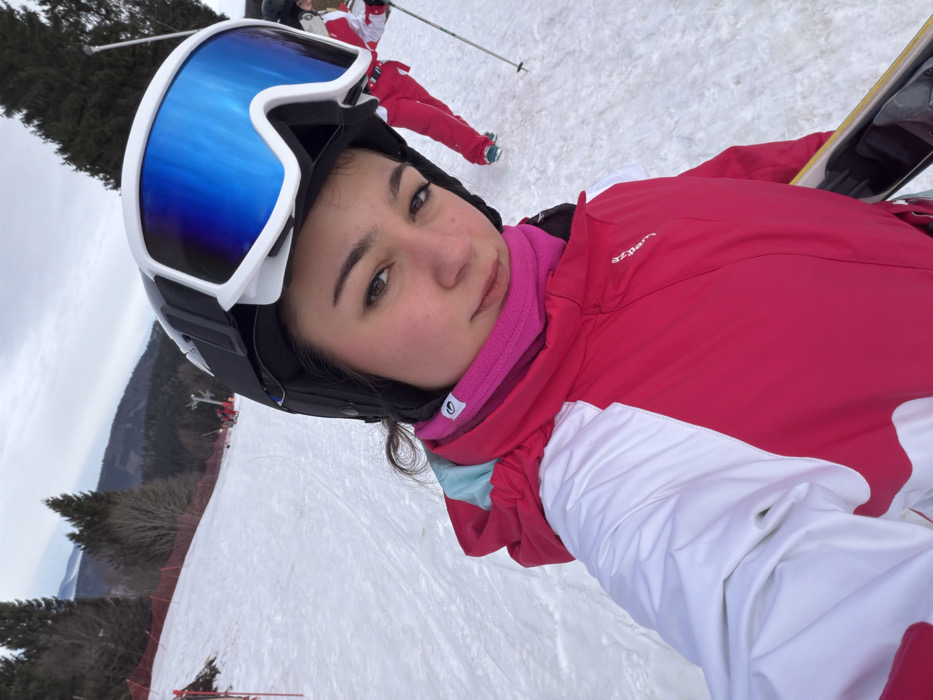 The image displays a young female on a snowy ski slope, identified as Belmont, France. She is positioned centrally, gazing directly forward. She wears a black ski helmet, white ski goggles with reflective blue lenses, a pink fleece neck gaiter, and a fuchsia and white ski jacket. Her right arm (viewer's left) is partially visible, holding a snowboard with a black binding. The background features a snow-covered mountain landscape under an overcast sky. A red safety net runs along the left side of the slope. Evergreen and bare deciduous trees are scattered across the hills and distant mountains. Another individual, wearing red ski pants and holding ski poles, is partially visible behind and to the right of the main subject. A ski lift tower is discernible further up the slope.