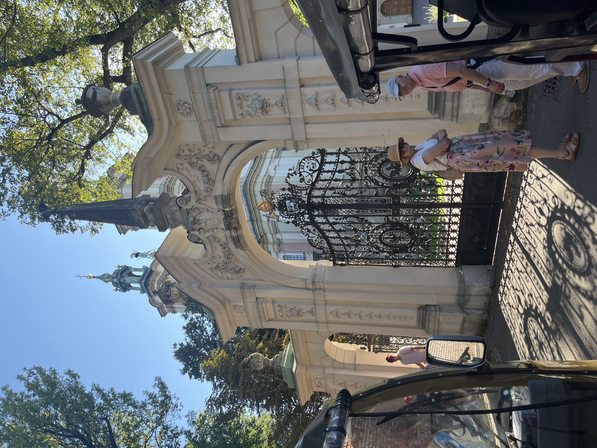 The image depicts a sunny day at the Wawel Royal Castle in Krakow, Poland, specifically in front of its ornate gate. Two individuals, a woman and a man, stand near the gate. The woman wears a straw hat, sunglasses, a white top, and a floral print skirt with sandals. She has her arms crossed. The man wears a light blue cap, a pink polo shirt, white pants, and casual shoes. He holds a bottle of water in his right hand. In the foreground, a portion of a golf cart or similar electric vehicle is visible on the left, and another on the right. The gate itself is made of intricate black wrought iron, featuring decorative circular motifs. Behind the gate, a light-colored building with arched windows and a domed structure with a cross can be seen. Lush green trees with dappled sunlight filtering through their leaves provide a natural backdrop, casting intricate shadows on the paved ground. The overall scene suggests a leisurely visit to a historic landmark.