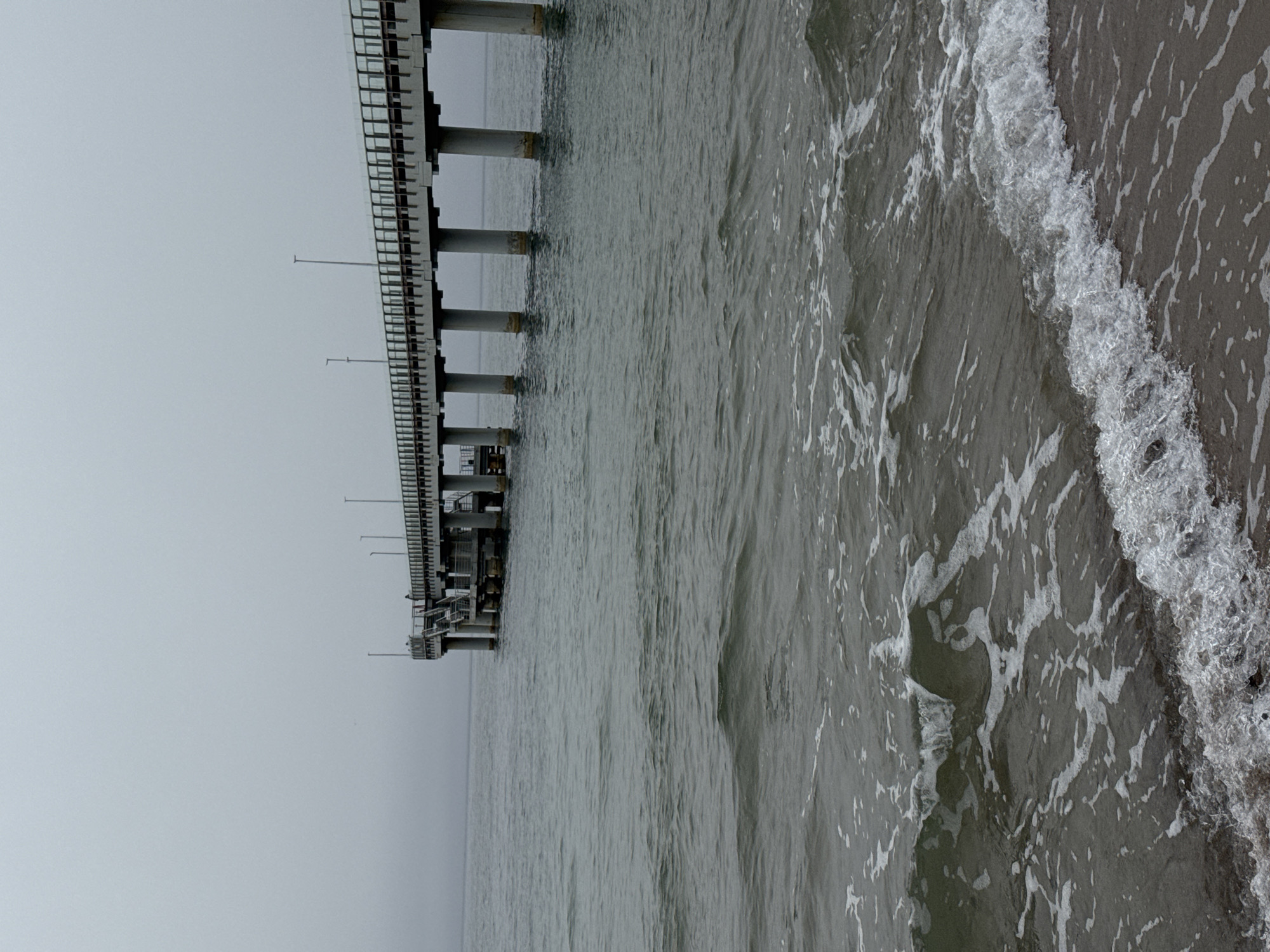 The image depicts a pier extending into the Baltic Sea on a gray, overcast day in Zelenogradsk, Russia. Waves are breaking on the beach to the right, with the gray sea stretching to the horizon. The pier is constructed of gray pillars and supports, with metal railings and what appears to be a platform at its end. There are a few light poles on the pier. The sky is a uniform gray color, blending into the sea in the distance.
