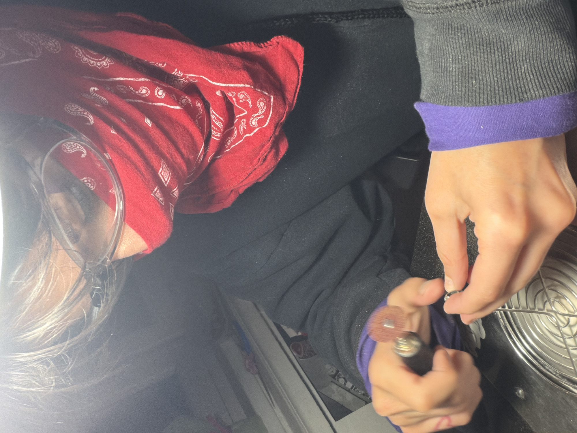 A person wearing safety glasses and a red bandana is using a rotary tool to work on a small metal object. The person is wearing a dark gray sweatshirt with purple cuffs and black pants. The object being worked on is placed on a metal grill, possibly a fan cover. The location of the image is not discernible. The city of Sofia, Bulgaria cannot be confirmed based solely on the provided visual information.